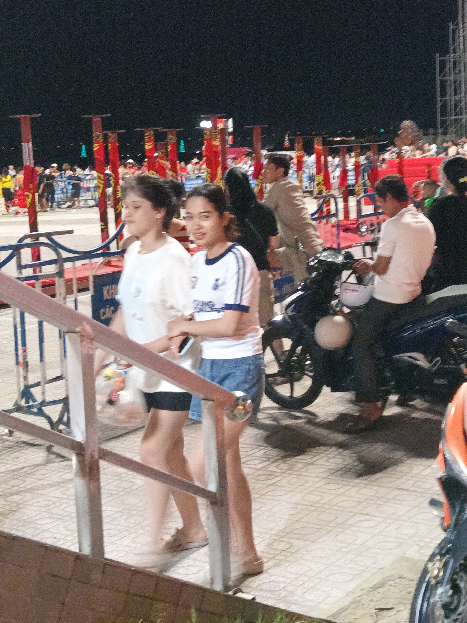 In Nha Trang, Vietnam, at night, two young women are walking on a tiled walkway. One woman, wearing a white t-shirt and dark shorts, is walking slightly ahead, holding a small bag of items. The second woman, in a white and blue striped t-shirt and denim shorts, is walking behind and to the side of the first, looking towards the camera with a smile. She is holding a smartphone in her right hand. Behind them, a man is sitting on a dark blue motorcycle, holding a white helmet. In the background, a crowd of people is visible, with red and gold banners and posts, suggesting a festive or event atmosphere. The walkway is bordered by a metal railing and fence.