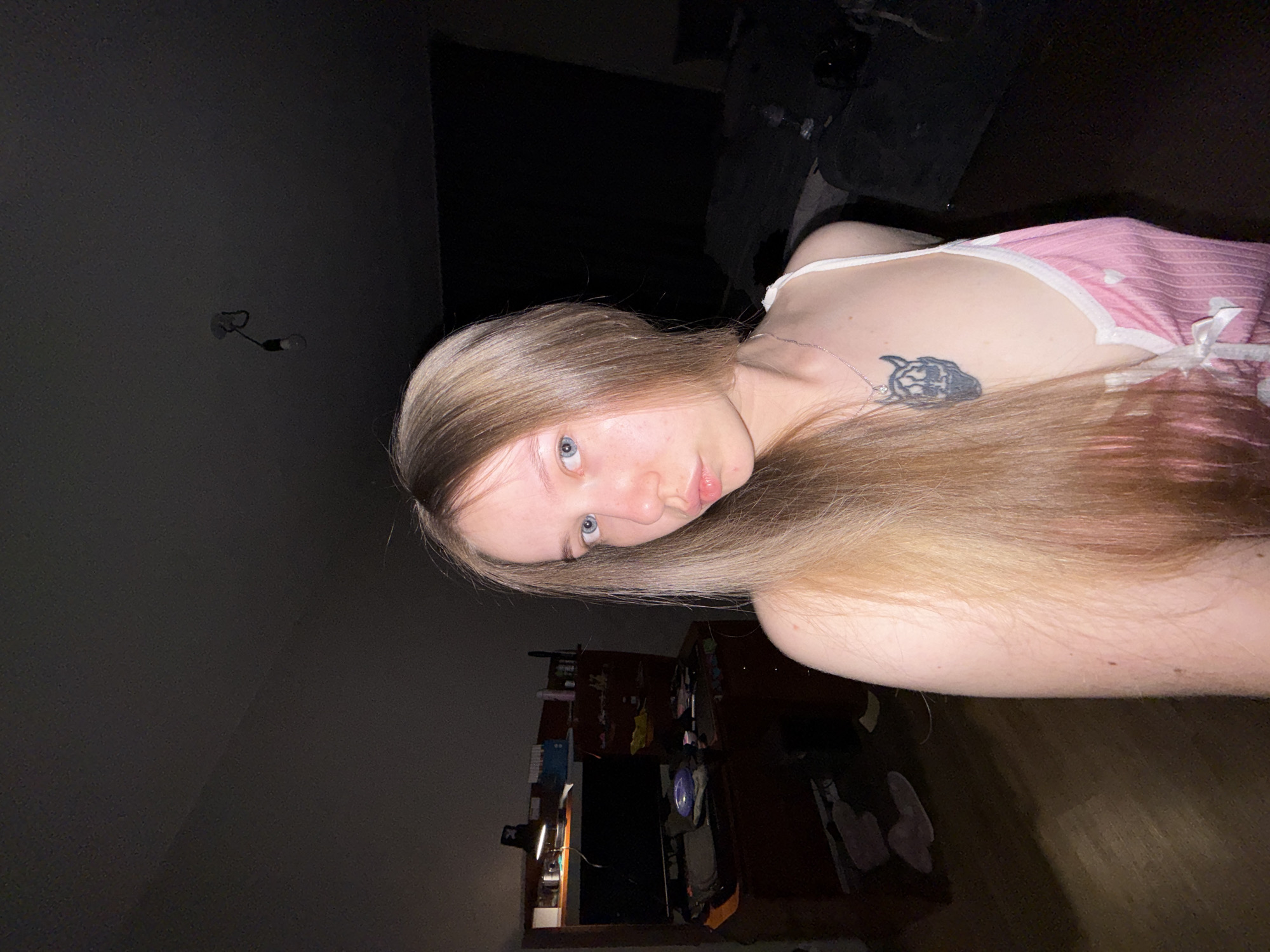 A young person with long, light brown hair and blue eyes is lying down, facing the camera. They are wearing a pink and white top. A tattoo is visible on their upper chest near the left shoulder. The background is dimly lit, showing a wall with a light fixture, a partially visible cluttered shelf unit with various items, and a dark-colored sofa or seating area. The location is likely indoors and given the user's location is in Chaikovsky, Russia, this is potentially a domestic setting in that city.