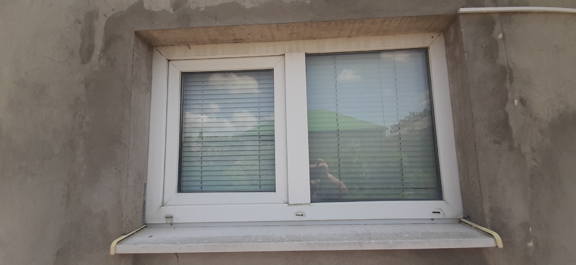 The image shows a close-up, eye-level exterior view of a white, double-paned sliding window installed in a textured gray concrete wall. The window has horizontal blinds, partially obscuring the view outside. In the reflection on the right pane, a person's head and shoulders are visible, holding a phone, suggesting they are taking the photograph. A green-roofed structure is reflected in the window as well. The wall appears to be under construction or renovation, with patches of unfinished concrete and stucco. A light-colored windowsill extends from the bottom of the window. The scene is likely an exterior wall of a building in Yessentukskaya, Russia, given the context.