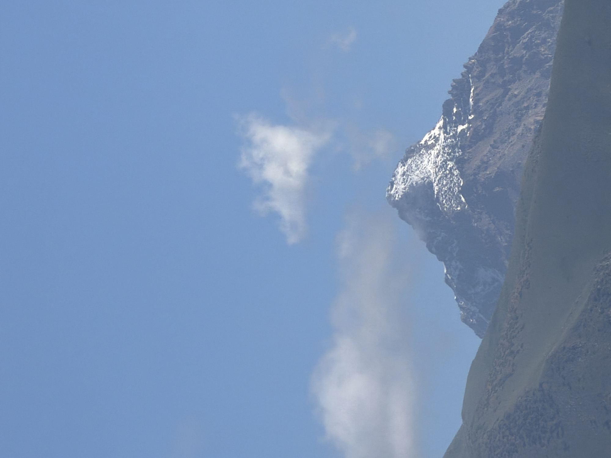 A snow-capped mountain peak rises from behind a dark, grassy mountain slope under a clear blue sky with a few wispy white clouds. The mountain is rugged with visible rocky outcrops and patches of snow clinging to its slopes. The foreground shows the textured, dark green and brown slope of a lower mountain, with subtle undulations. The lighting suggests it is daytime, with shadows indicating the sun is likely to the right of the frame.