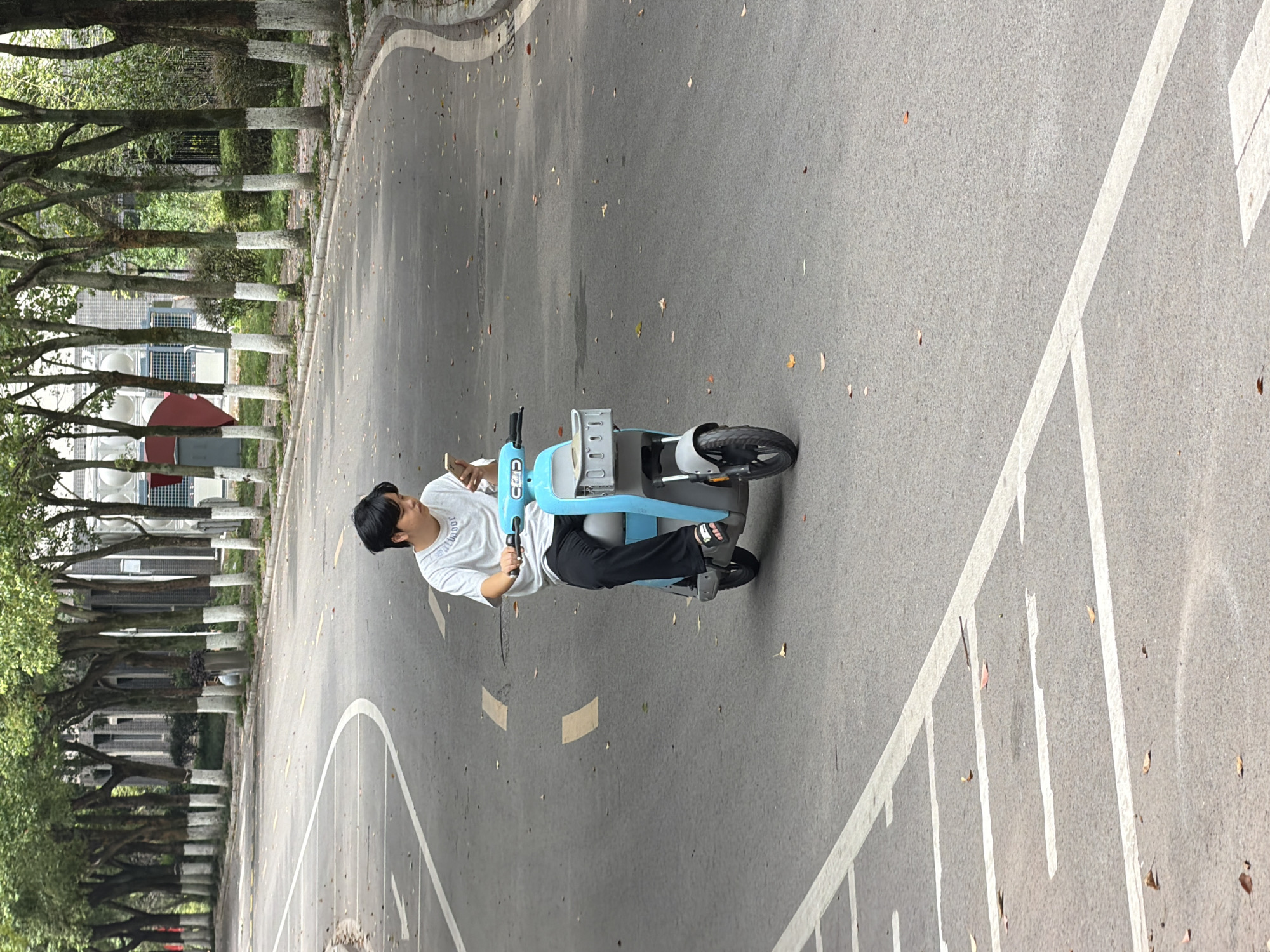 A young East Asian male wearing a white t-shirt and black pants is riding a light blue electric scooter on a paved road. He is holding a smartphone in his right hand and looking down at it. The scooter has a grey basket attached to the front. The road has white lane markings, and fallen leaves are scattered on the pavement. In the background, there are several trees lining the road, with some buildings visible behind them. The image appears to have been taken outdoors on a cloudy day. The location is the city of Jiangjin, China.