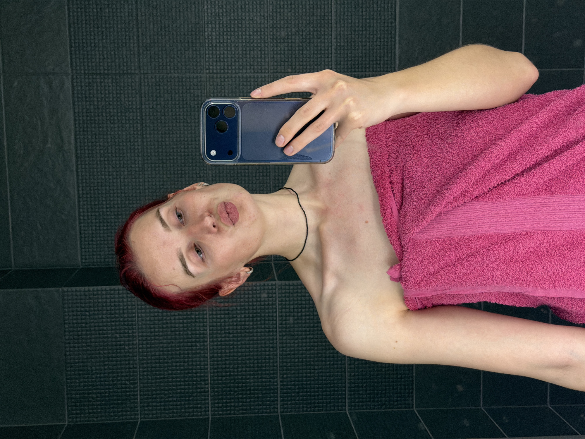 A young woman with red hair pulled back is taking a selfie in a bathroom. She is wearing a pink towel wrapped around her body and a thin black choker necklace. She is holding a dark blue smartphone in her right hand and making a kissing face towards the camera. The background consists of dark grey textured tiles. The image appears to have been taken in a residential bathroom. The city of Chaikovsky, Russia, is not visible or implied in this image.