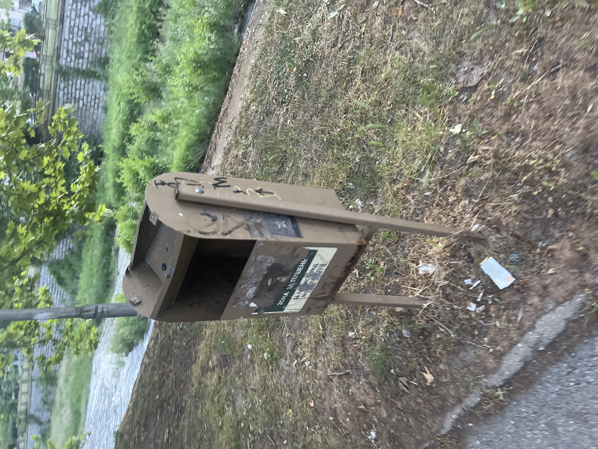 An outdoor scene shows a brown, weathered metal trash receptacle affixed to a metal pole and base. The receptacle has graffiti on the lid and a sticker on its front. The receptacle is surrounded by overgrown grass, debris, and a partially visible sidewalk. The location is likely in Blagoevgrad, Bulgaria, based on the Cyrillic text on the sticker on the receptacle. No people are visible in the image.