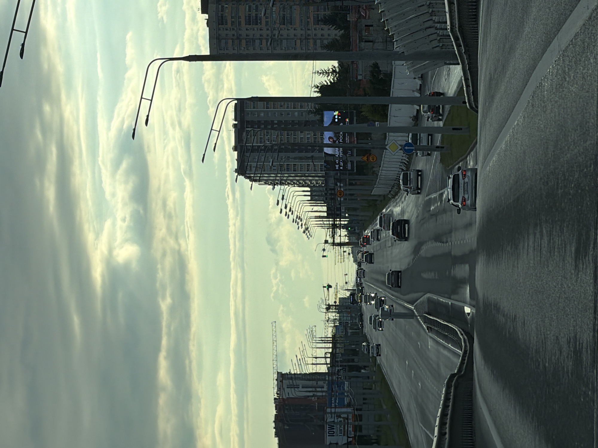 The image shows a highway in an urban environment. The sky is overcast with clouds, and the sun is low, casting a diffused light. The highway is wet, indicating recent rain or mist. Cars are driving on the highway in both directions. On the left side of the highway, there are several tall buildings under construction, with cranes visible. There are also streetlights and billboards along the road. On the right side of the highway, there is a barrier and more streetlights. The scene appears to be during the daytime, but the lighting suggests it might be early morning or late afternoon.
