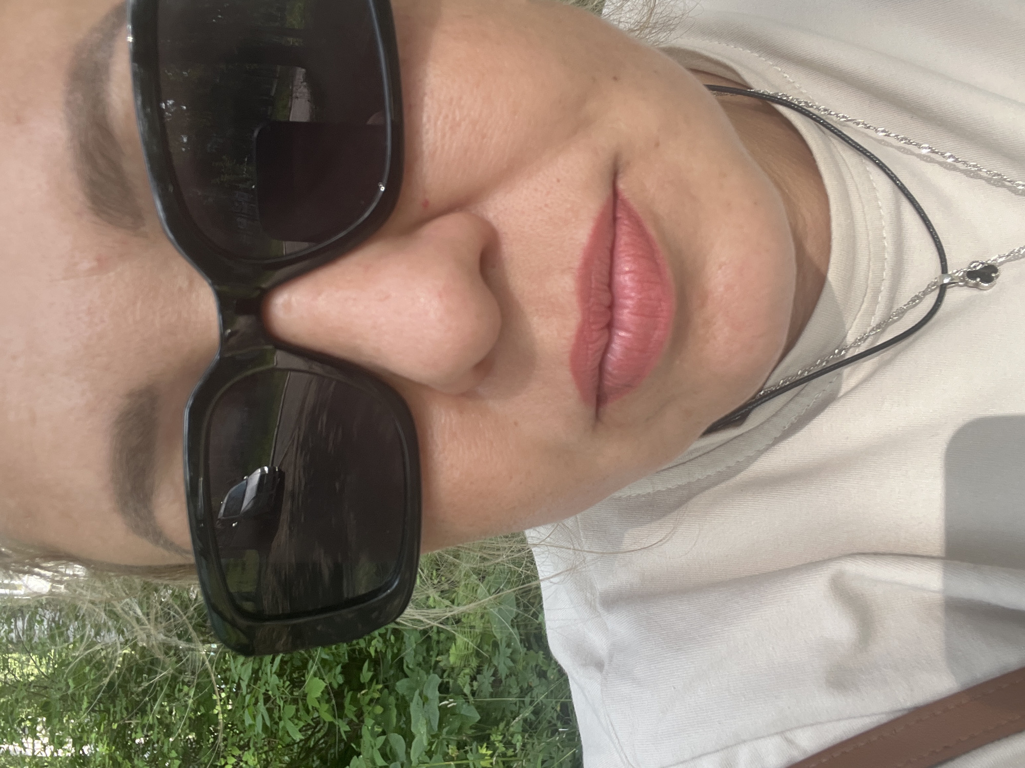 A medium shot, eye-level selfie captures a woman's face and upper torso, with a soft focus background. She is wearing large, black-framed sunglasses, which reflect a dark-colored van driving on a paved road. Her blonde hair is visible around her temples and pulled back. She has light skin with some visible pores and a few faint freckles. Her eyebrows are dark and well-defined, and she is wearing light pink lipstick. A light-colored t-shirt is visible, along with two necklaces, one black cord and one silver chain with a clover-shaped pendant. A brown strap from a bag is visible on her left shoulder. The background consists of lush green foliage, suggesting an outdoor, possibly park-like setting. No specific identifiable landmarks indicate the location in Chaikovsky, Russia.