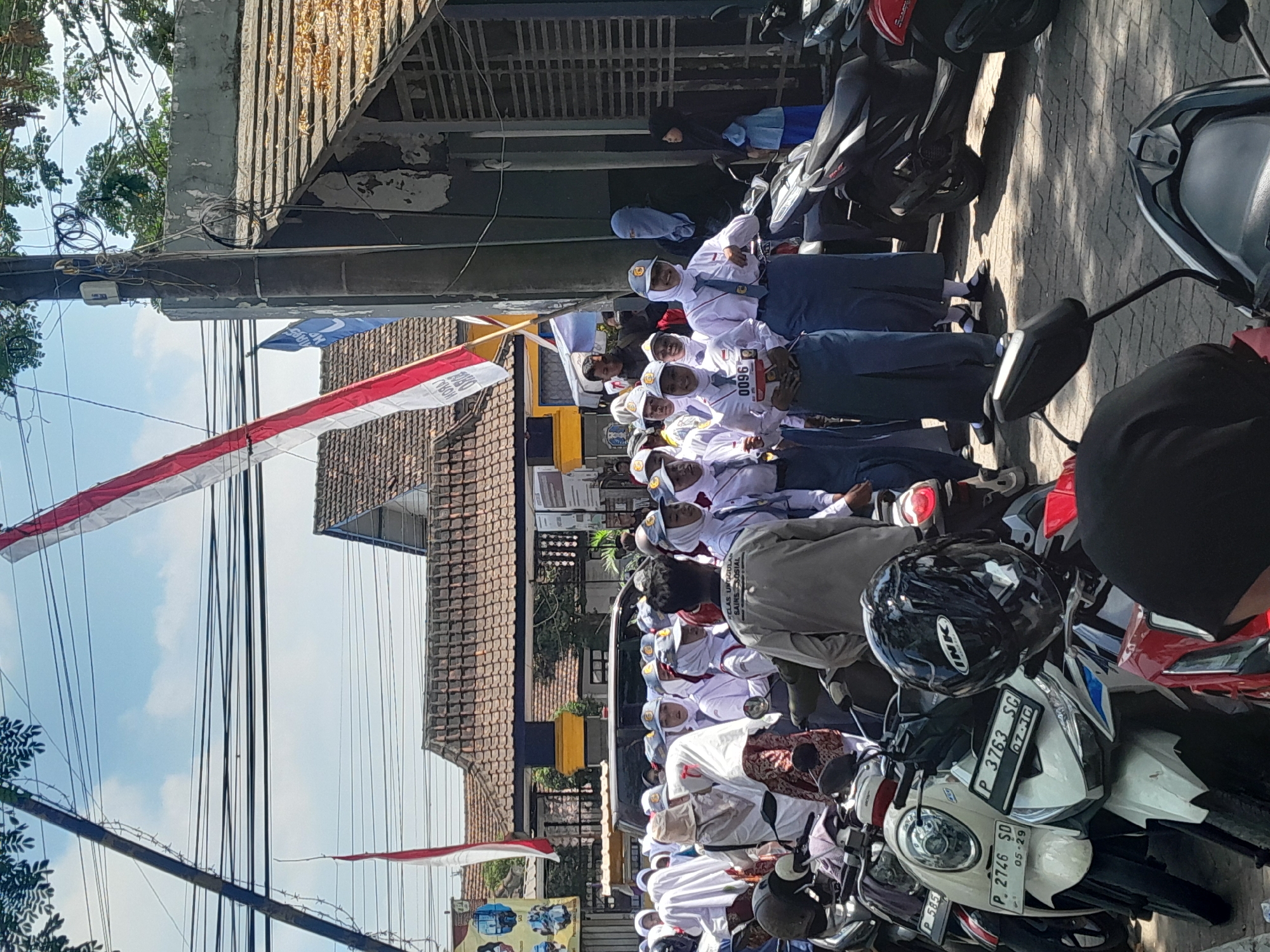 The image depicts a scene in Genteng, Indonesia, likely during a school event or parade. A group of students, predominantly female, are dressed in white shirts, blue skirts, and hats, with some wearing hijabs. Several students are carrying or wearing numbers, suggesting a competition or organized activity. The students are standing in a street lined with parked motorcycles, indicating a common mode of transportation in the area. In the background, there are buildings with tiled roofs and utility poles with numerous wires, characteristic of urban infrastructure. Two Indonesian flags are visible, one large red and white flag suspended diagonally across the upper portion of the frame, and another smaller red and white flag on a pole to the left. The environment appears to be outdoors on a sunny day.