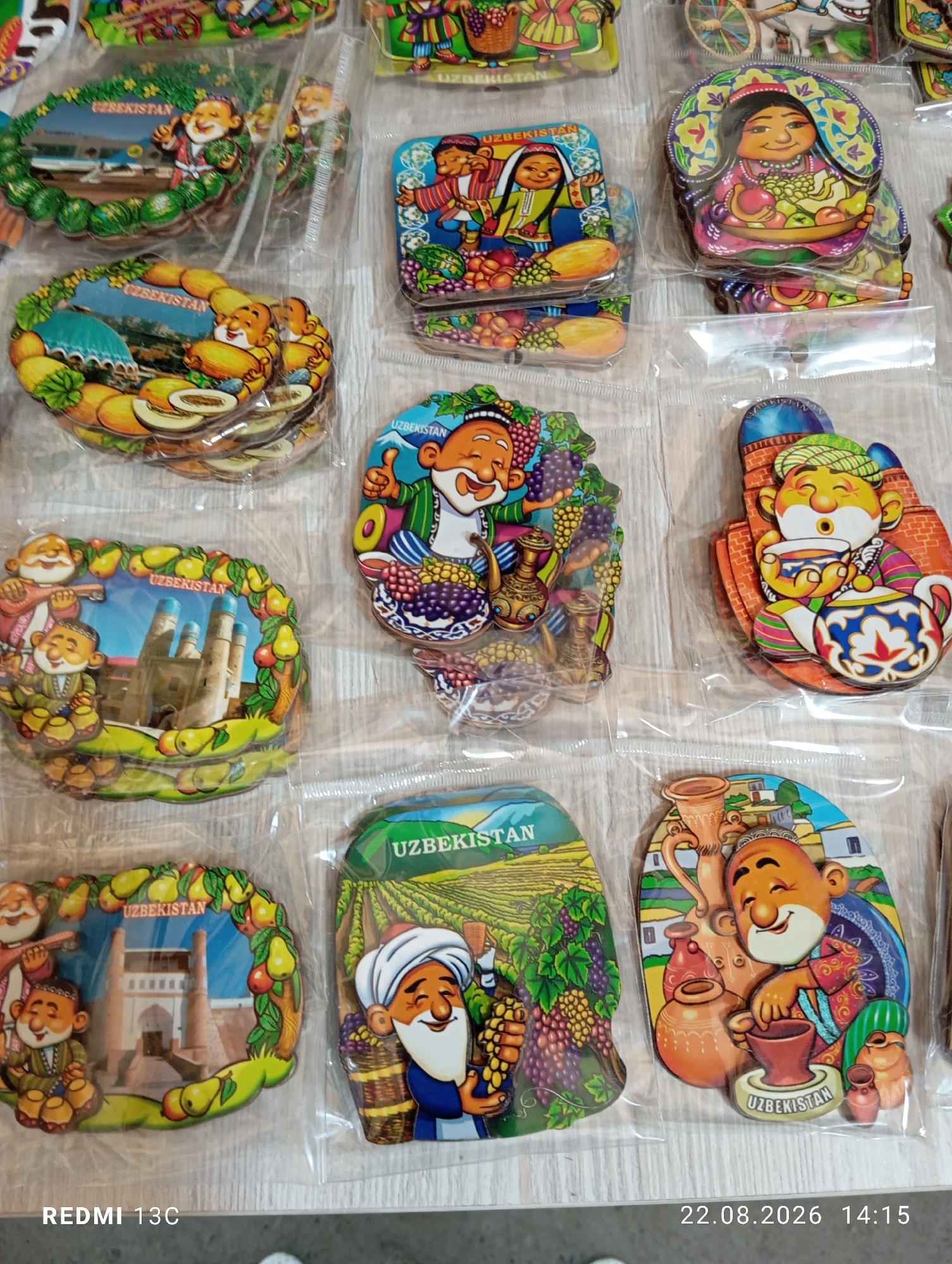 The image displays a collection of souvenir magnets, likely from Uzbekistan, laid out on a flat surface. The magnets feature cartoonish illustrations of people, often depicted in traditional attire, engaged in various activities such as playing music, holding fruit, or drinking tea. Many of the magnets also incorporate cultural landmarks, such as minarets, and images of local produce like grapes and melons. Text on some magnets clearly states "UZBEKISTAN". The scene is a close-up shot, suggesting the souvenirs are being displayed or sold. There is no discernible indication of the city of Amirsay, Uzbekistan, within the visual content. The lighting appears to be overhead, casting subtle shadows.