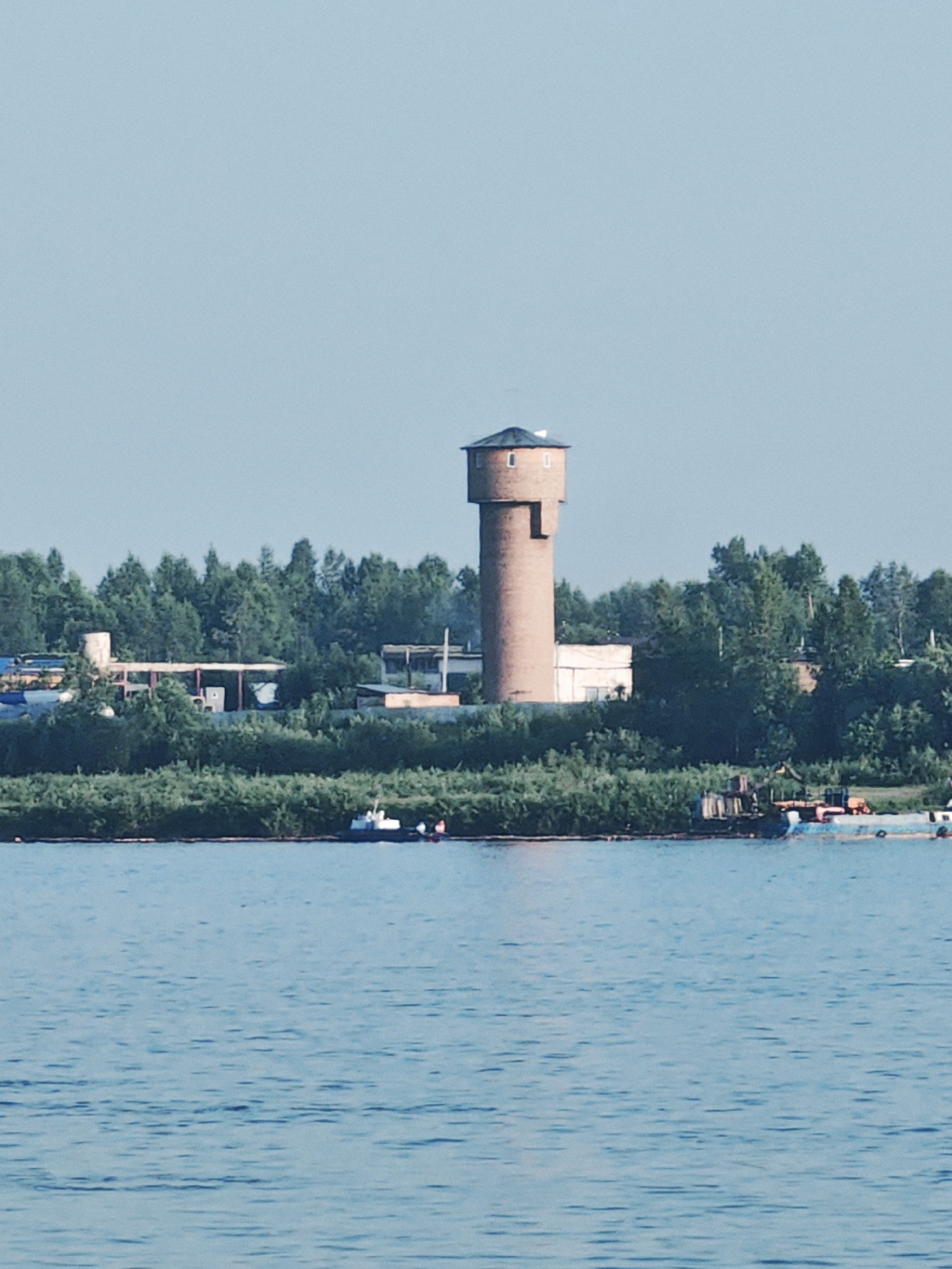 A long-range, vertical, full-shot captures a brick water tower on the outskirts of Lesosibirsk, Russia. The tower stands prominently against a clear, pale blue sky, surrounded by a dense line of green trees. In front of the trees, a body of calm, blue water stretches across the bottom of the frame. A few boats are visible on the water near the shore, and some industrial buildings are partially obscured by the trees behind the tower. The scene suggests a somewhat rural or industrial area bordering a body of water.