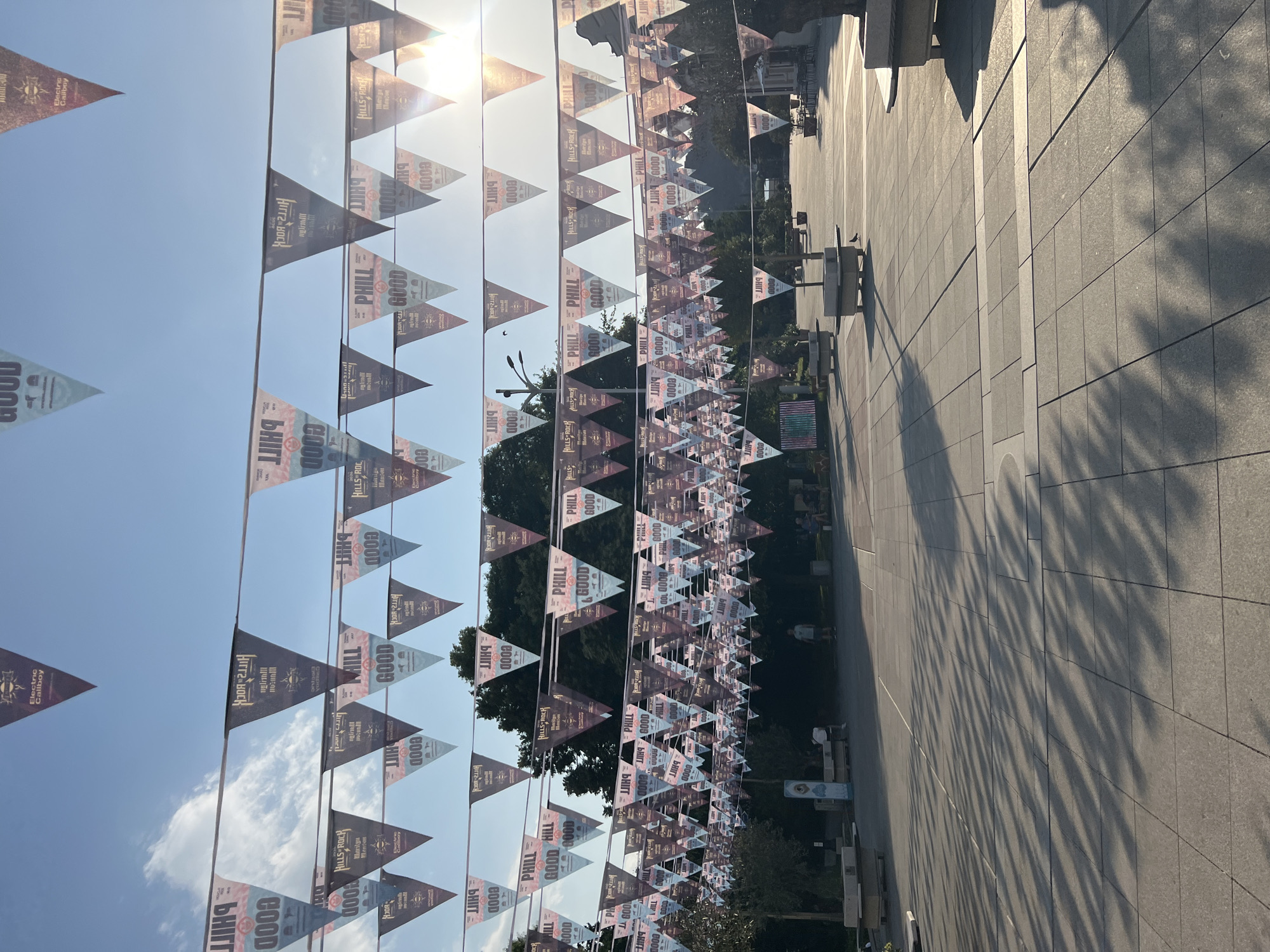 The image depicts an outdoor public space, likely a square or pedestrian area, adorned with numerous triangular pennant flags strung across wires. The flags are predominantly dark with white and red lettering, featuring text that appears to be "PHILL GOOD" and "HILLS ROCK". The setting is illuminated by bright sunlight, casting long shadows from trees and possibly structures onto the paved ground. In the background, trees, a lamppost, and some buildings are visible. Several people are present in the scene, some sitting on benches and others walking. The ground is paved with large square tiles. The overall impression is of a festive or event-related decoration in an urban environment. This scene is in the Kapana district of Plovdiv, Bulgaria.