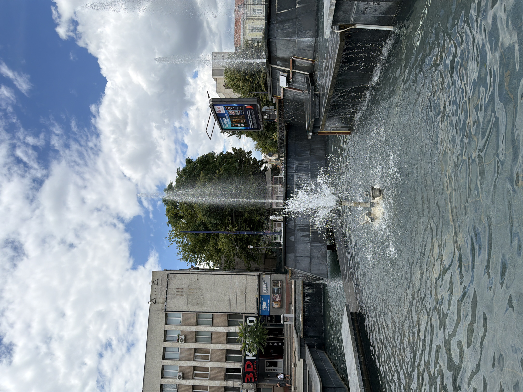 The image depicts a public fountain in the city of Pazardzhik, Bulgaria. The fountain features multiple jets of water spraying upwards and cascading down tiered, dark-tiled structures. The water is clear and rippling, reflecting the cloudy sky above.
In the background, a multi-story building with a light-colored facade and numerous windows is visible. Signs on this building indicate a "3D CINEMA" and the political party "GERB". Adjacent to this building, a large billboard displays advertisements, with "A1" prominently featured. Lush green trees surround the buildings and fountain area, suggesting a pleasant, possibly urban park setting.
The sky is a mix of blue sky and white, fluffy clouds, indicating fair weather. The overall scene is one of an active public space, likely a town square or plaza, with the fountain serving as a central element. No people are clearly identifiable in the foreground, but a few figures can be seen near the entrance of the cinema building in the background. The architecture of the buildings suggests a Central or Eastern European aesthetic.