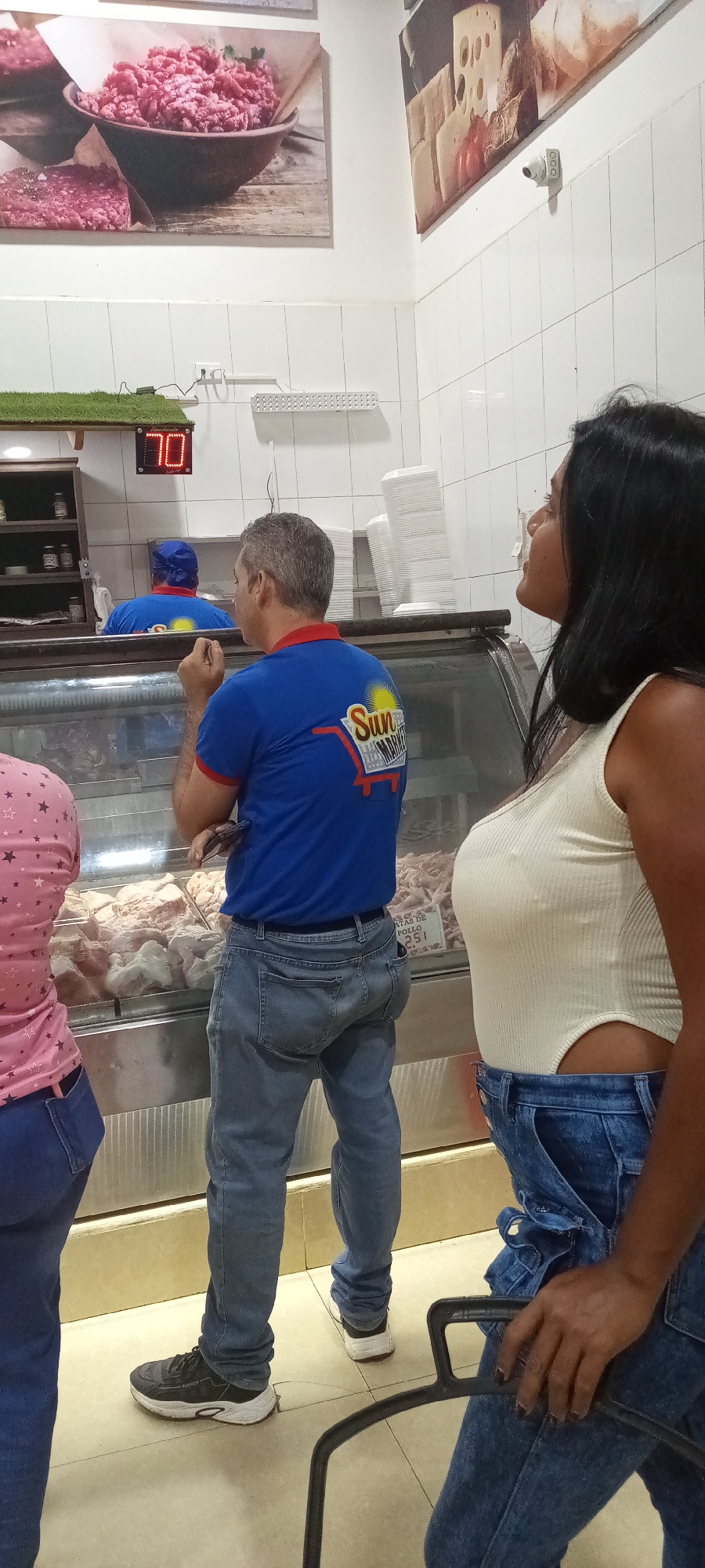 The image shows three customers at a meat counter in what appears to be a butcher shop or a supermarket. The location is Unknown, Unknown. A man in a blue shirt with a "Sun Market" logo on the back is looking at the meat selection inside the glass display case. To his left, partially visible, is another customer wearing a pink shirt. To the right of the man, a woman with long black hair is standing, also looking at the meat display. She is wearing a white tank top and denim overalls tied around her waist. In the background, behind the counter, a worker wearing a blue uniform and cap can be seen. Above the worker, there is a digital display showing the number 70. On the wall above the counter, there are framed pictures of meat and cheese. A security camera is mounted on the wall near the cheese picture. The floor is tiled, and a portion of a shopping cart is visible on the right.