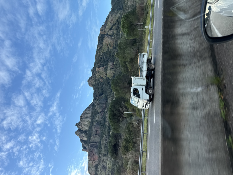 A white flatbed truck travels on a highway alongside a mountain. The mountain has a rocky face with sparse tree cover. A blue sky with scattered clouds is visible. The view is from inside a vehicle, with a side mirror in the upper right corner, reflecting the scenery. A concrete median runs along the right edge of the road.