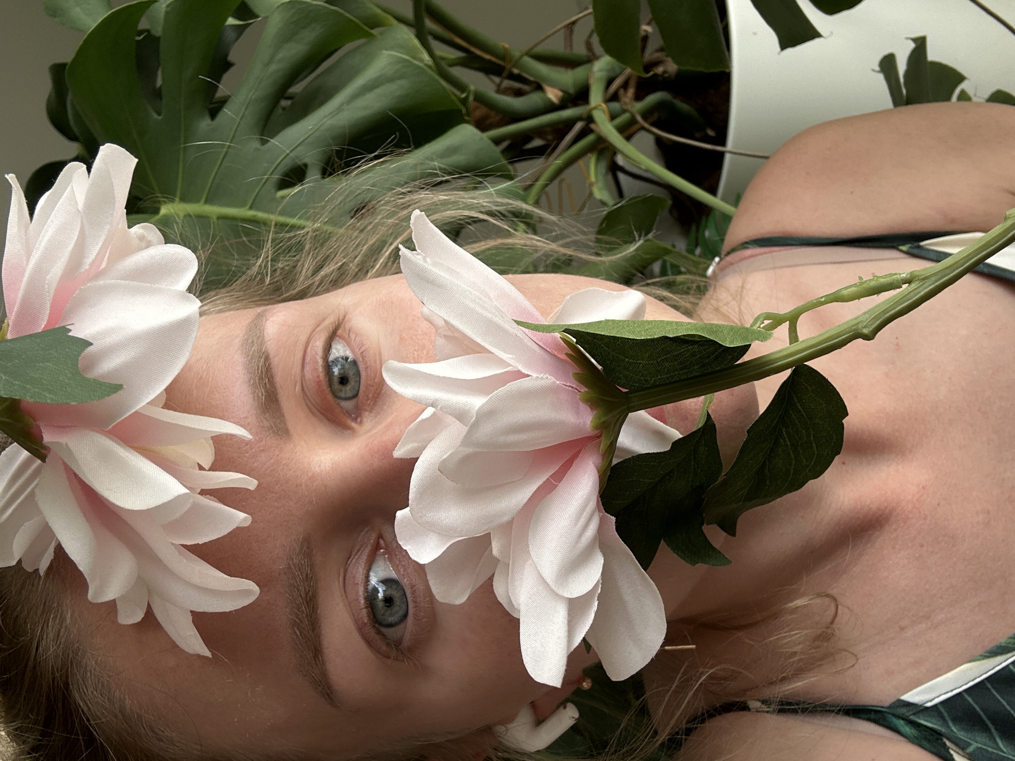 The image is a close-up, portrait-oriented shot of a person lying down amidst foliage and flowers. The person has light skin, blonde hair, and blue eyes. A large white and light pink flower with green leaves is positioned near the right eye and cheek, and another similar flower is near the forehead on the left side of the frame. Green Monstera leaves are visible in the background, along with some leafy vines. The person's body is oriented sideways, with a glimpse of the shoulder and upper chest area, where a dark green and white patterned garment is visible. The overall composition creates a natural, somewhat whimsical, and intimate scene. The location is unknown.