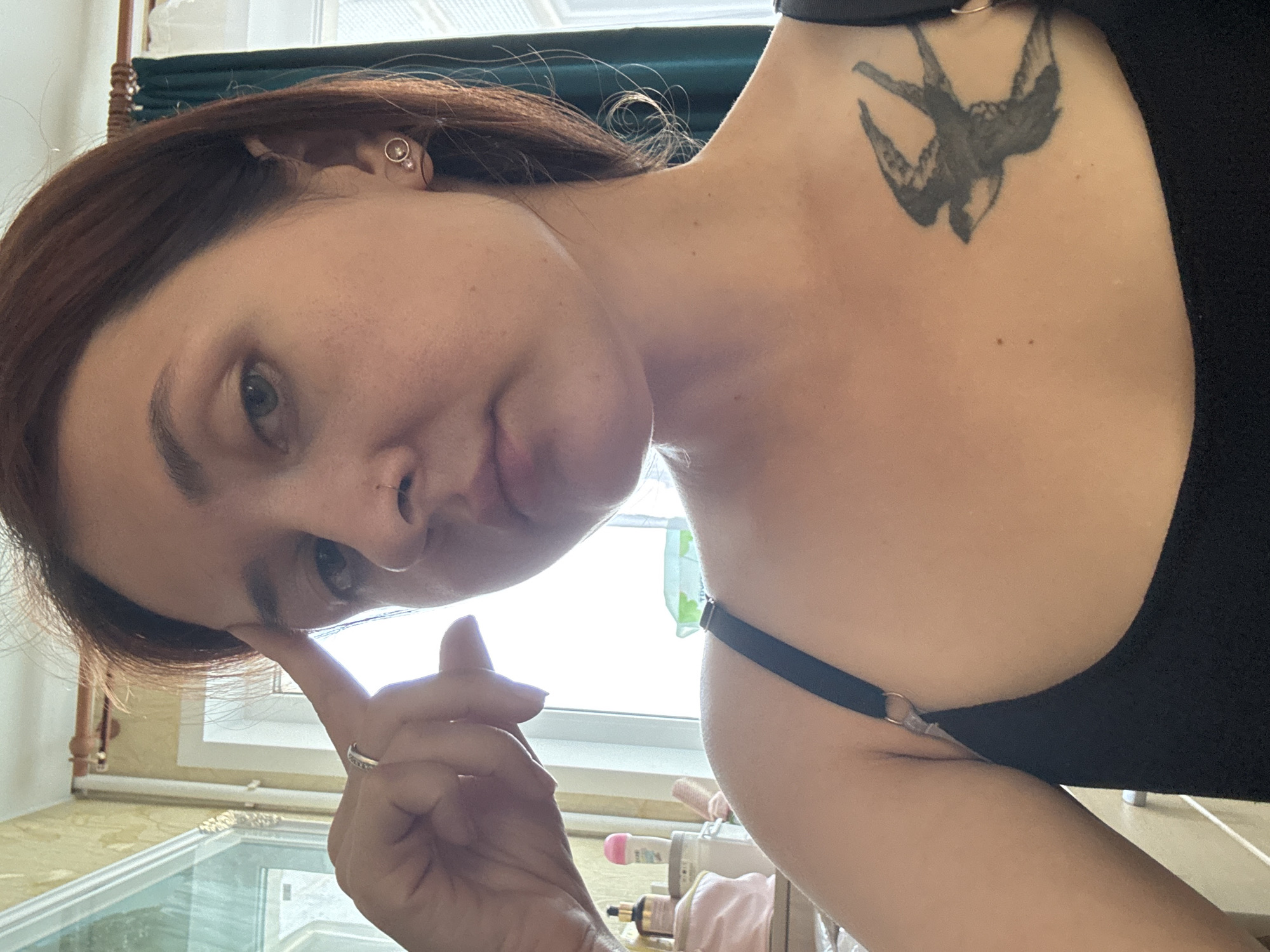 A close-up, selfie-style shot shows a woman with medium-length, reddish-brown hair and a light complexion. She is wearing a black tank top, revealing a tattoo of a swallow on her upper chest. She has a nose ring and a ring on her left ring finger. Her right hand is raised to her forehead. In the background, there is a window with sheer curtains, a radiator, and some toiletries on a shelf. The scene appears to be indoors, possibly a home, and the lighting suggests it is daytime. There is no discernible information about the location in Naberezhnye Chelny, Russia.