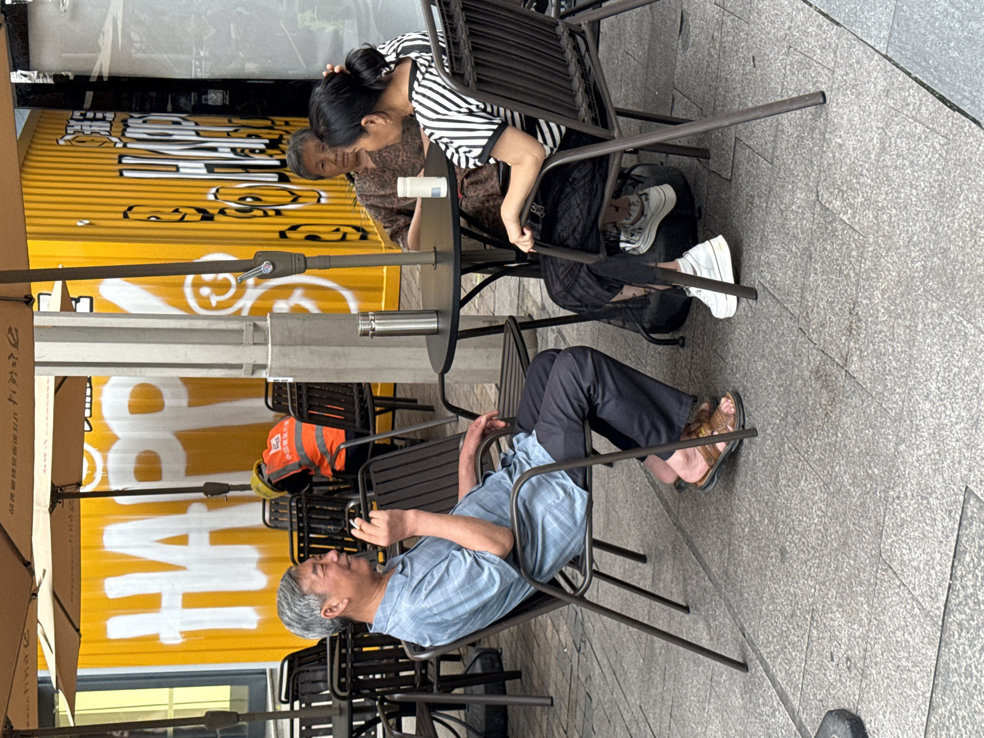 Here is a description of the image:
An outdoor urban scene shows three people sitting at a metal table and chairs on a tiled surface, possibly a public square or patio. The location is suggested to be in the city of Jiangjin, China. An older man with grey hair wearing a blue shirt and dark pants is seated in a chair, appearing to hold a cigarette in his hand. Across the table, an older woman wearing a patterned brown shirt and younger woman in a striped black and white shirt are seated. They both face the same direction as the man. Behind them is a bright yellow structure with white and black graphics, appearing to be either a building or a large advertisement. The scene is illuminated by natural light.