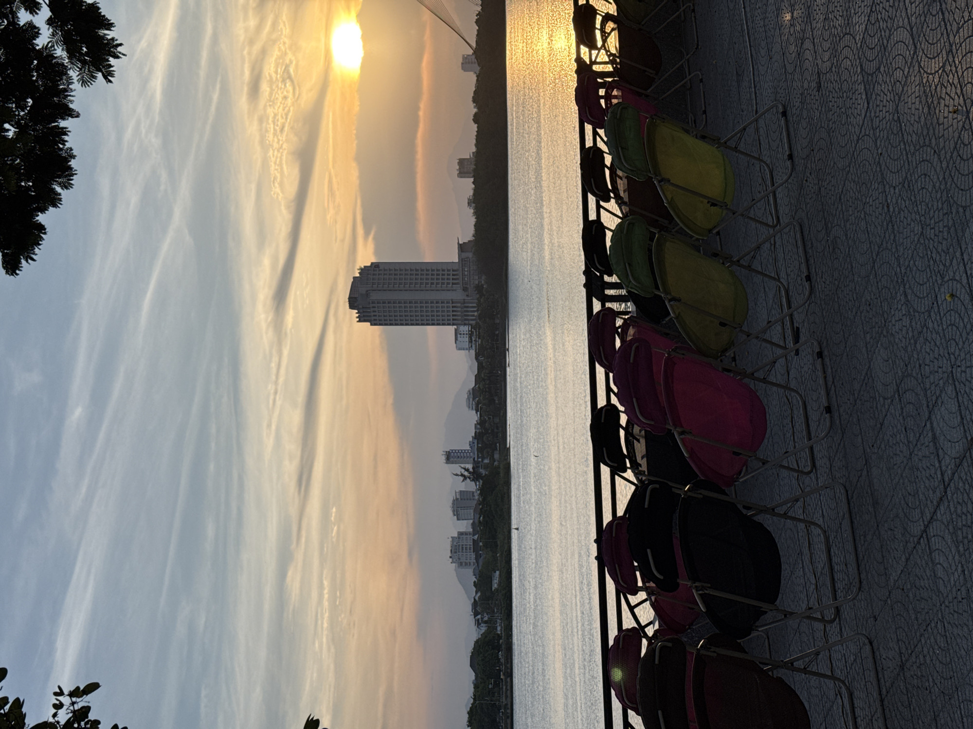 In Đà Nẵng, Vietnam, a row of empty folding chairs faces a wide body of water. The chairs are arranged on a paved area with a decorative tile pattern. Beyond the water, a city skyline is visible, dominated by a tall, modern building. The sun is setting on the horizon, casting a warm, golden glow across the sky and water, with streaks of orange and yellow clouds. Silhouettes of trees frame the top of the image. The scene appears to be an outdoor seating area for an event or gathering, set against a scenic backdrop of the city and the sunset.