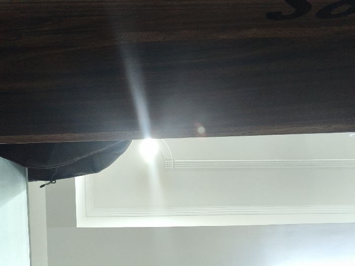 The image shows a portion of a ceiling in a room, likely a living room or bedroom, located in Gwalior, India. The ceiling is white, with an ornate molding design and a single white light fixture visible. The image is taken from an angle, looking up at the ceiling from beneath a dark brown wooden structure, likely a beam or ledge. The wooden structure has a faint "S" shaped carving on its surface. The lighting in the room is bright, casting a glare on the surface of the wooden structure. There are no people or other subjects visible in the image.