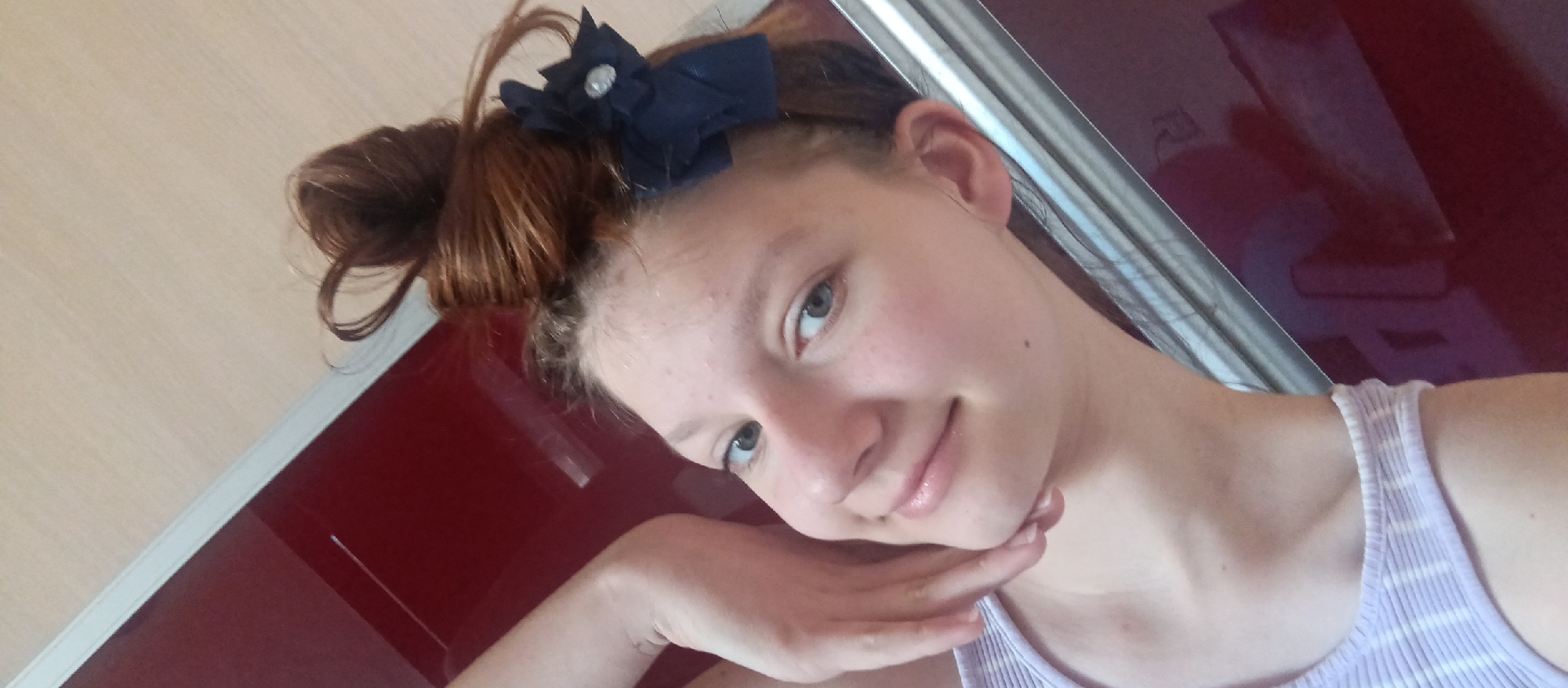 A young woman with reddish-brown hair styled in a messy bun adorned with a dark blue bow is positioned in front of a burgundy-colored surface with white trim, possibly a door or cabinet. She is wearing a light-colored tank top with subtle stripes. The woman is tilting her head and smiling gently, her chin resting on her left hand. The background appears to be indoors, with a light-colored wall partially visible behind the burgundy surface. There is no readily discernible information to indicate a specific location within the city of Unknown, Unknown.