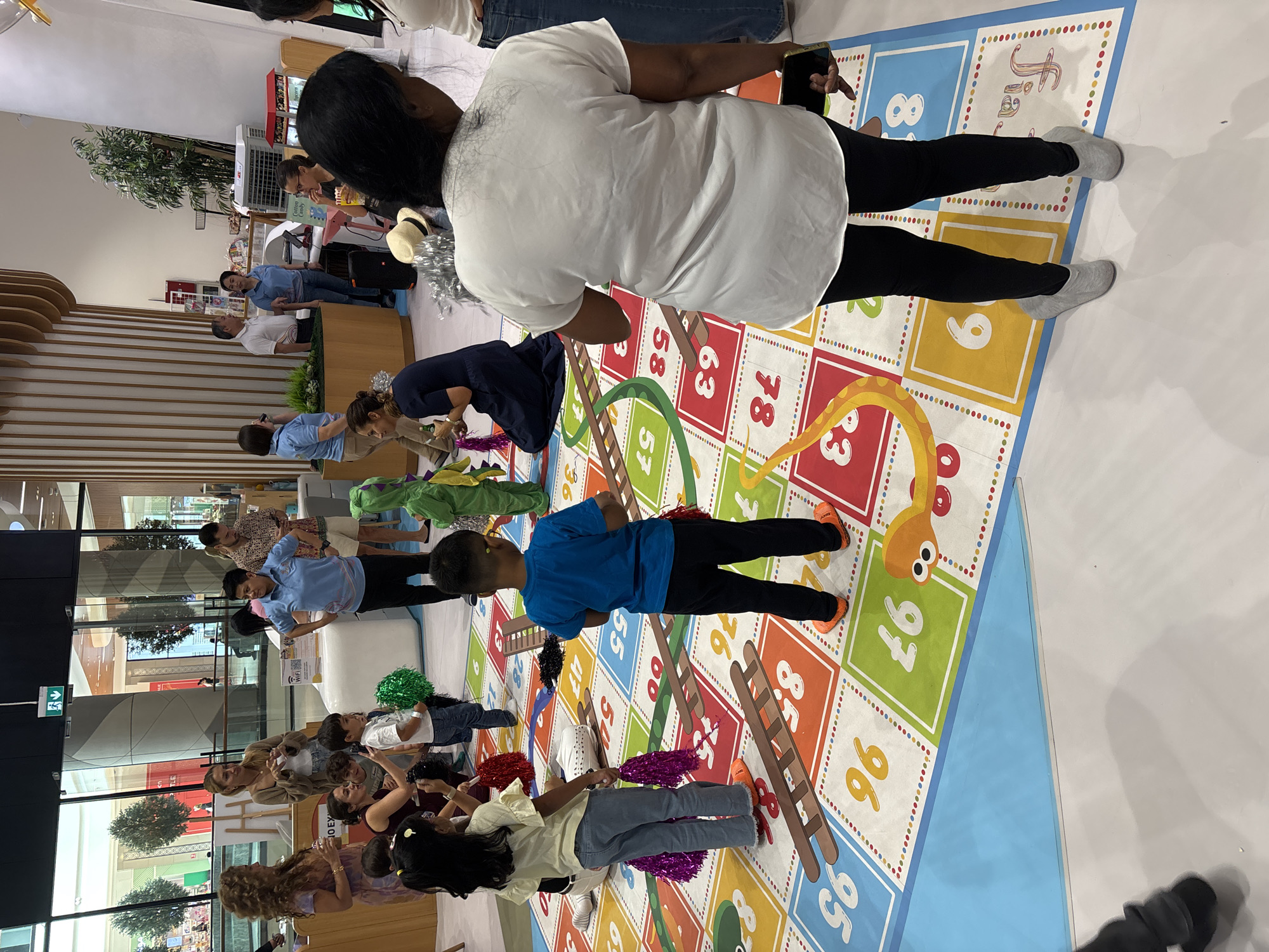 The image depicts an indoor scene, likely a public space or event venue in Dubai, United Arab Emirates, where a large, colorful floor game resembling Snakes and Ladders is set up. Several individuals, including adults and children, are present, interacting with the game and each other.
In the foreground, two children are standing on the game board. One child, wearing a blue t-shirt and black pants, is facing away from the camera, positioned on a square with the number 97. Another child, a girl with a yellow shirt and blue jeans, is facing towards the game board, holding purple pom-poms. She is standing on a square marked with the number 45. A third child, dressed in a green dinosaur costume, is standing near the top right of the game board, facing left.
Several adults are also visible. A woman in a white top and black leggings is standing on the right side of the image, holding a phone in her right hand. She is wearing grey slip-on shoes. Another woman in a dark blue dress is crouched on the left side of the game board, holding a green pom-pom.
In the background, more people are gathered, some standing and observing, while others appear to be staff members. There are architectural elements typical of a modern indoor public space, including large windows, pillars, and wooden paneling. A white counter or booth is visible behind some of the adults. The overall atmosphere suggests a playful and engaging event.