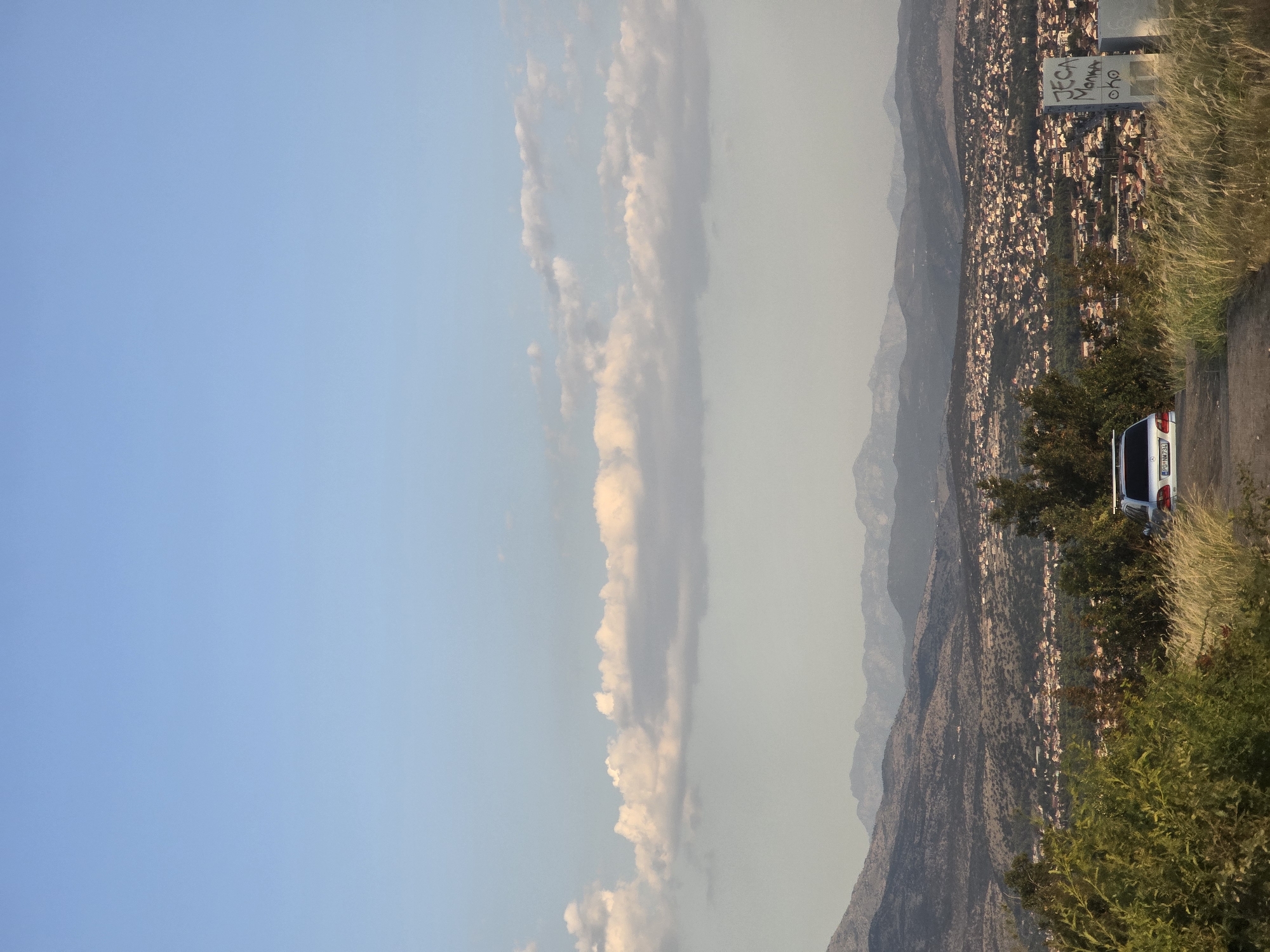 The image depicts a panoramic view of the city of Podgorica, Montenegro, from a vantage point on a hill. The sky is a clear blue with scattered white clouds. In the foreground, lush green vegetation and tall, dry grass line a dirt road. A silver sedan is parked on the right side of the road.
The middle ground reveals a sprawling urban landscape with numerous buildings, predominantly with red-tiled roofs, nestled amongst rolling hills. The city extends towards the horizon, where a range of majestic mountains forms a dramatic backdrop. The atmosphere is serene, with the soft light suggesting either early morning or late afternoon.
On the far right of the image, a concrete structure with graffiti is visible. The graffiti appears to be in Cyrillic script and is difficult to decipher entirely, but some readable words include "JECA," "MONKA," and "OHO." The overall scene captures the characteristic blend of urban development and natural beauty that defines Podgorica's landscape.