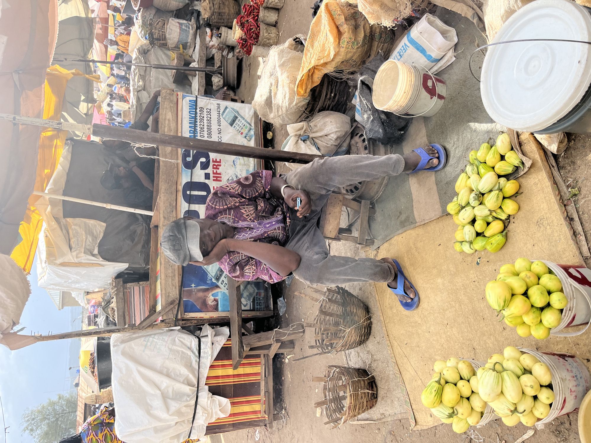 The media file depicts a market scene in Rafin Par, Nigeria. The primary subject is a person resting in a shaded area, leaning against a wooden structure. They are wearing a purple patterned shirt and grey pants, with blue flip-flops on their feet. The person appears to be asleep or resting their eyes, with one hand near their face and the other resting on their lap.
The setting is an outdoor market with various stalls and goods. Prominently displayed are several buckets filled with light green and yellow striped produce, likely vegetables. Piles of what appear to be dried goods and sacks are also visible in the background. Woven baskets are scattered around, some empty and others containing items. The ground is covered with a light-colored mat or cloth.
In the background, other market activities can be inferred, with glimpses of people and more stalls under awnings. The lighting suggests it is daytime, likely with bright sunlight outside the shaded areas. The overall atmosphere appears to be that of a busy, working market.
Visible text includes a sign with Nigerian phone numbers and what appears to be a business name like "ADABANKOWA". Another sign shows a calculator and the text "HFRE". The general environment suggests a traditional market setting.