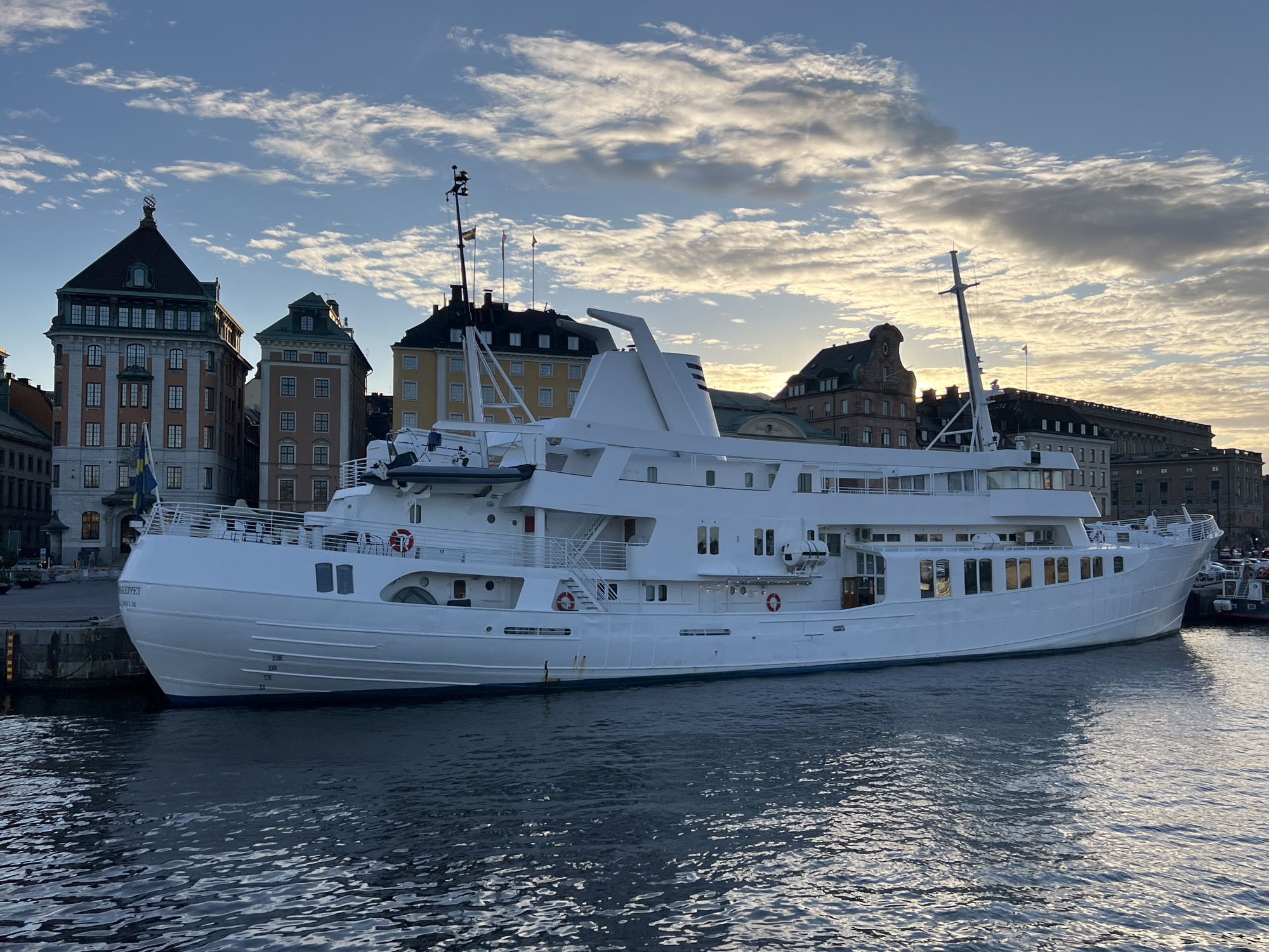 An exterior, eye-level, medium-wide shot captures a large, white motor yacht moored in the Stockholm harbor, with a backdrop of historic buildings and a cloudy sky during the day. The yacht is the primary subject, occupying the majority of the frame, with its multiple decks, railings, and windows visible. The water in the foreground is dark and rippled, reflecting the sky and the yacht. Behind the yacht, several multi-story buildings with distinctive architecture stand along the waterfront, appearing to be from different eras. The sky is a mix of blue and white clouds, with sunlight breaking through, suggesting either sunrise or sunset. A Swedish flag is visible on a mast on the yacht. The scene appears tranquil and urban, showcasing a blend of maritime activity and city life in Stockholm.