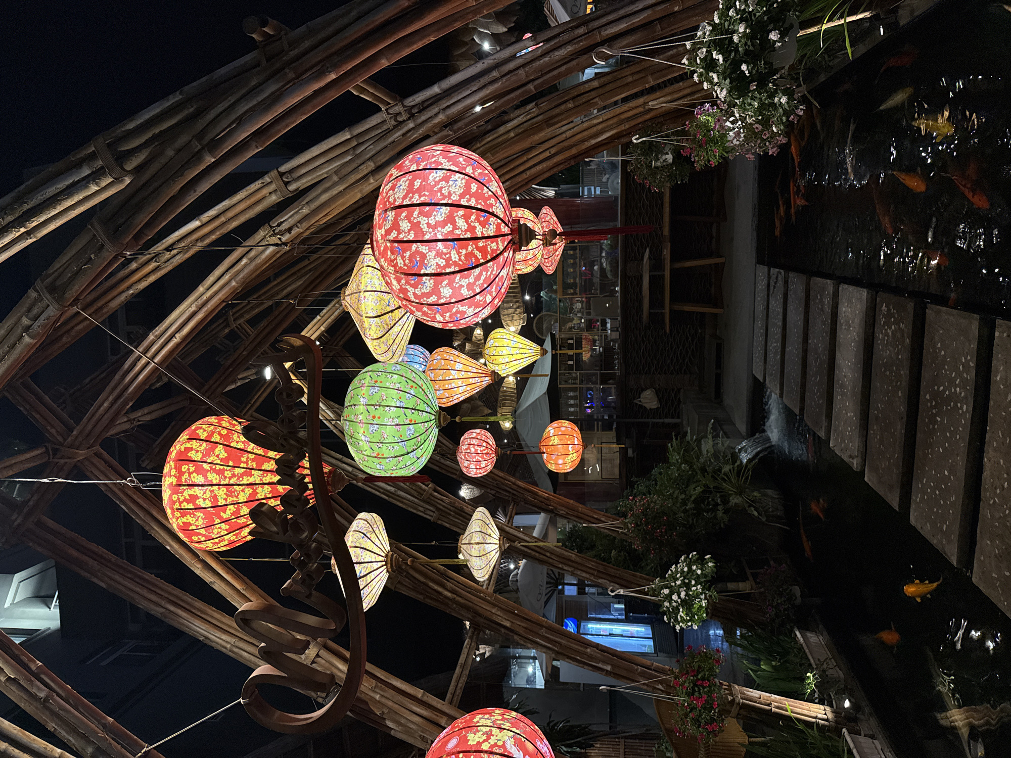 This night-time image captures a beautifully lit outdoor setting, likely a restaurant or garden area in Nha Trang, Vietnam. The scene is dominated by an elaborate structure crafted from bamboo poles, forming an arched or sloped canopy.
Numerous colorful lanterns hang from this bamboo framework, acting as the primary light source. These lanterns vary in shape, including large spherical ones and more elongated, traditional styles. They are adorned with intricate patterns; for instance, prominent red lanterns display white and blue floral designs, while green lanterns feature similar motifs, and yellow and orange lanterns show small yellow flowers. The glowing lanterns create a warm, inviting ambiance against the dark background.
To the right side of the frame, a dark body of water, clearly a koi pond, is visible. Several koi fish, appearing orange and yellow, swim in the water. Concrete or stone stepping stones line the edge of the pond. Hanging baskets filled with white and pink flowers are suspended above the pond and integrated into the bamboo structure, adding to the decorative appeal.
No people are visible, and no text is discernible in the image. The overall impression is one of tranquility and cultural aesthetics, characteristic of a pleasant evening setting.