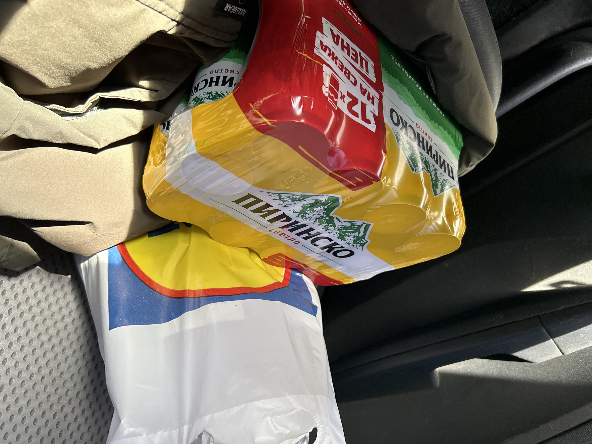 Here is a detailed description of the image:
The image shows items on a gray car seat. A khaki-colored "PULL&BEAR" jacket is partially visible on the left side of the frame. Next to it is a multi-pack of "ПИРИНСКО светло" beer (Pirinsco Svetlo) wrapped in clear plastic, its packaging showing the text "12 x HA CBEЖA ЦЕНА" (12 x FOR FRESH PRICE) and "свежо от планината" (fresh from the mountains). Beneath the beer pack, there is a white plastic bag with a yellow and blue design, which appears to be a shopping bag. The seat's gray fabric texture is visible. The scene takes place inside a car, with sections of the car's interior in the background.