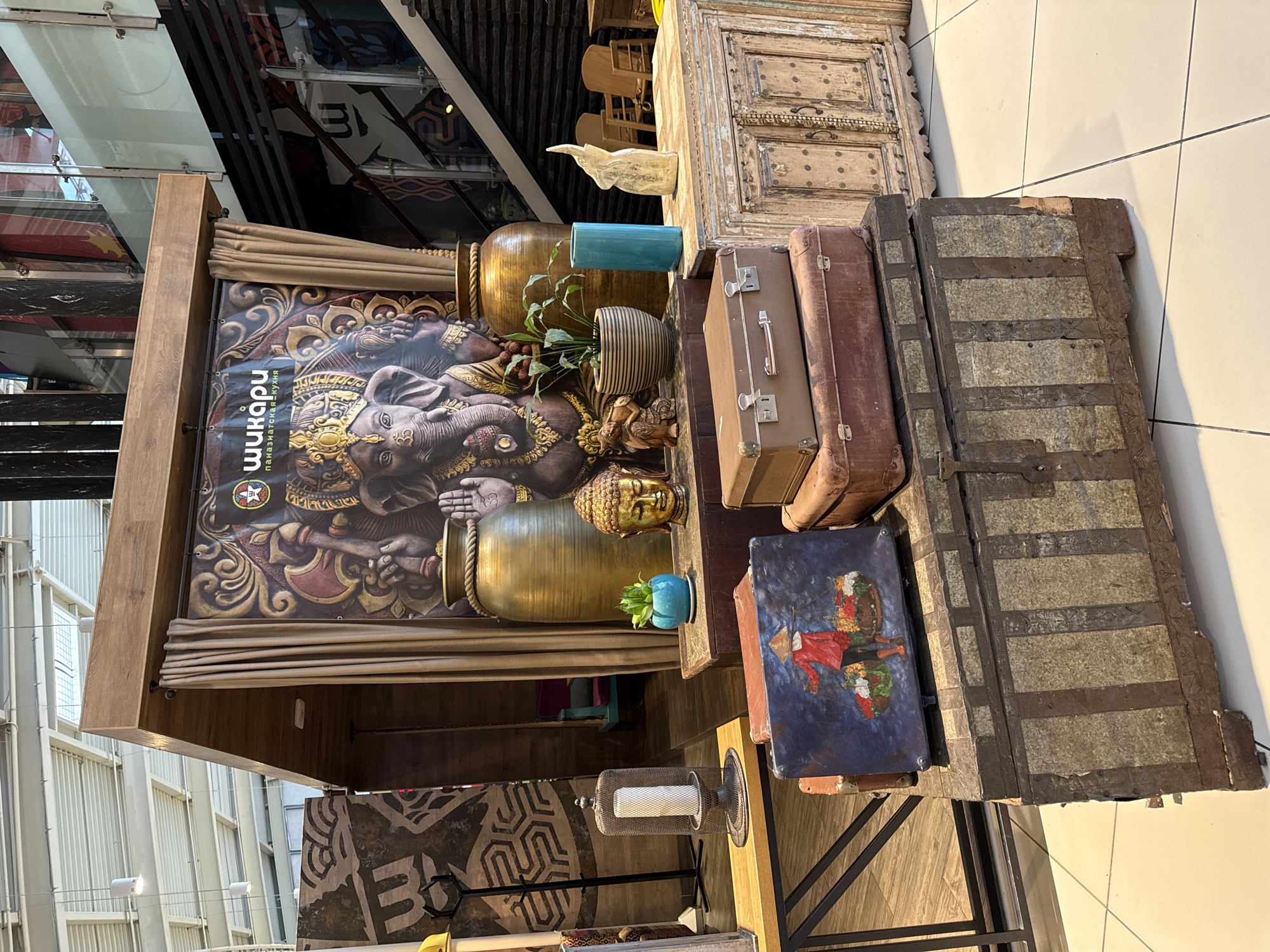 This indoor shot features a display of eclectic items stacked on and around a wooden structure. The scene is likely set in a commercial space, possibly a shopping mall, in Khimki, Russia, given the presence of a sign in Russian that reads "Шикари" (Shikari), a known restaurant in that city.
The display includes a large framed image of a Ganesha-like figure, antique-looking wooden cabinets and chests, suitcases, vases, and decorative objects like a golden Buddha head and a candle lantern. The items are arranged in a somewhat haphazard manner, suggesting a deliberate attempt to create a bohemian or vintage aesthetic. The lighting is bright and even, typical of a retail environment.
The environment appears clean and modern with tile flooring and glass or reflective surfaces visible in the background, indicating a contemporary architectural design. There is no immediate sign of people in the frame, leading focus to the arrangement and the objects themselves.