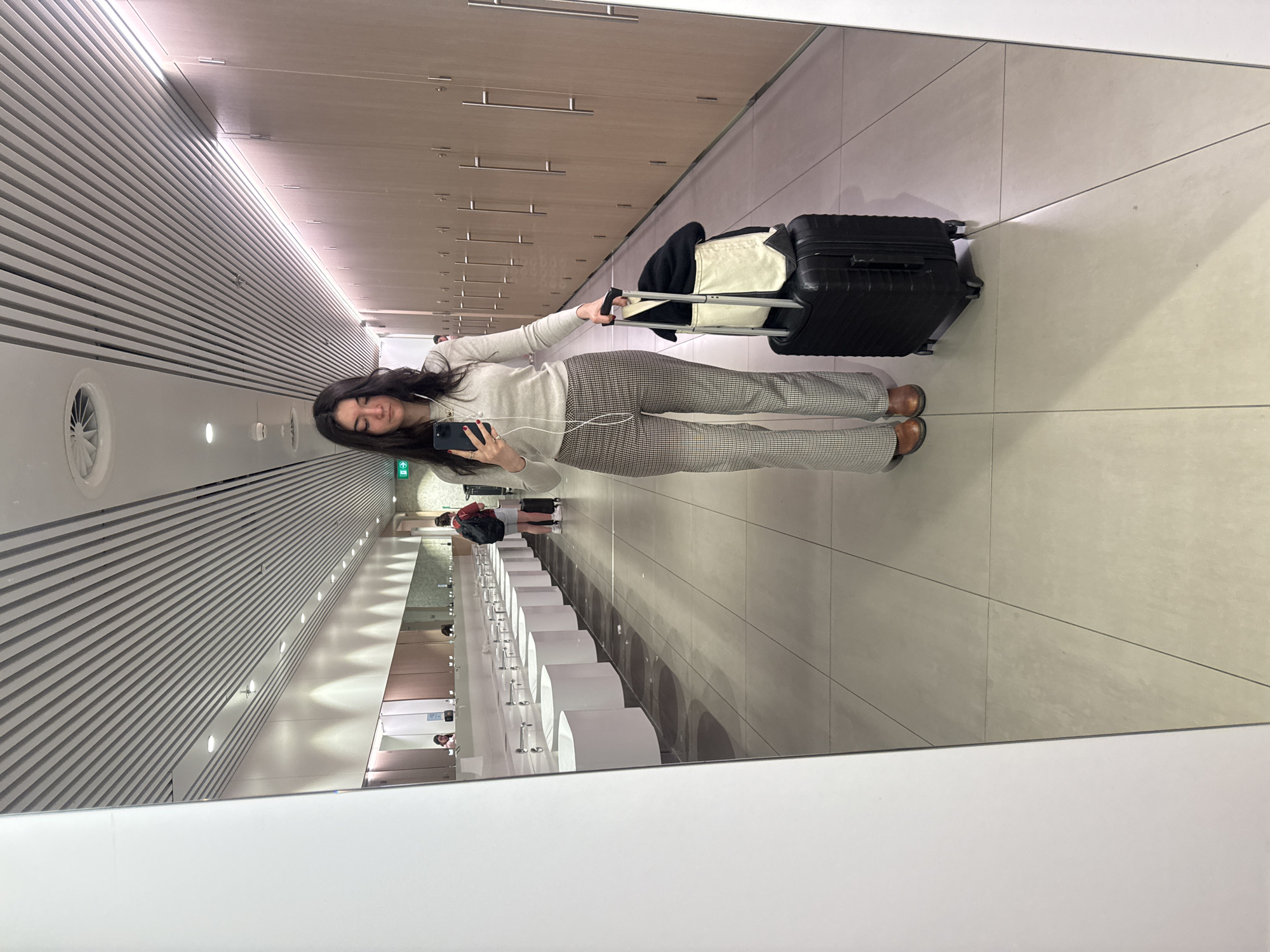 A mirror selfie shows a woman in an airport bathroom, likely in Schiphol, Netherlands. She wears a light gray sweater, patterned pants, and brown shoes, pulling a black suitcase with a white bag atop. The background reveals a modern airport bathroom with long rows of sinks and a hallway lined with wooden cabinets. Two other people are visible in the background. The mirror's edge is visible in the foreground, indicating a public space. The mirror reflects a long hallway with slatted walls and ceiling.