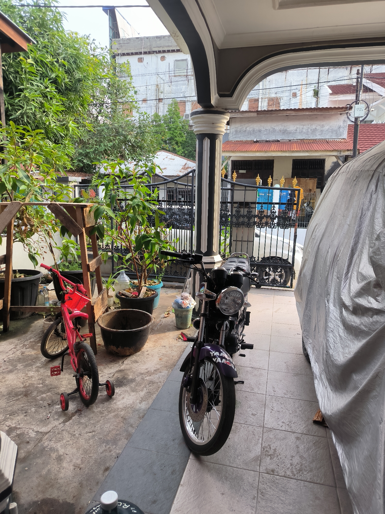 This image depicts a residential scene likely in Lhokseumawe, Indonesia, based on architectural style and potential local objects. A partially covered vehicle is visible on the right, next to a paved area where a motorcycle and a small red bicycle with training wheels are parked. To the left, there is a small garden or outdoor storage area with potted plants, wooden supports, and various containers. A black metal gate separates the immediate area from the street, revealing houses or buildings in the background. The scene appears to be in a residential neighborhood.