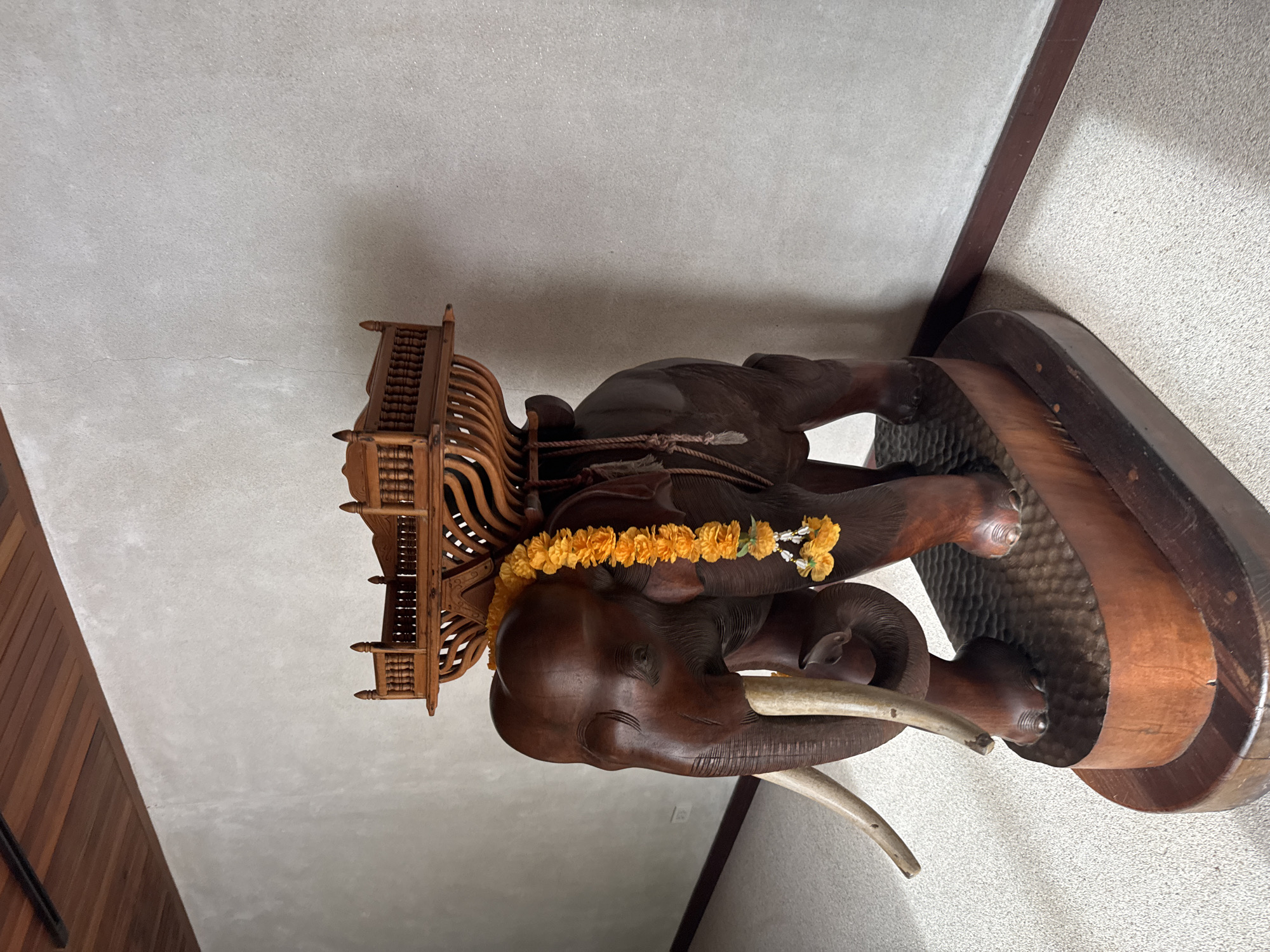A dark brown, intricately carved wooden elephant statue stands on a two-tiered wooden base. The elephant is adorned with a yellow marigold garland draped around its neck and smaller yellow floral ornaments hanging from it. On the elephant's back rests a decorative, multi-tiered wooden structure resembling a palanquin or howdah. The statue is positioned indoors, against a textured, light-colored wall. The floor is carpeted with a light beige, speckled material. The ceiling features dark wooden planks. The overall setting appears to be a hallway or interior space.