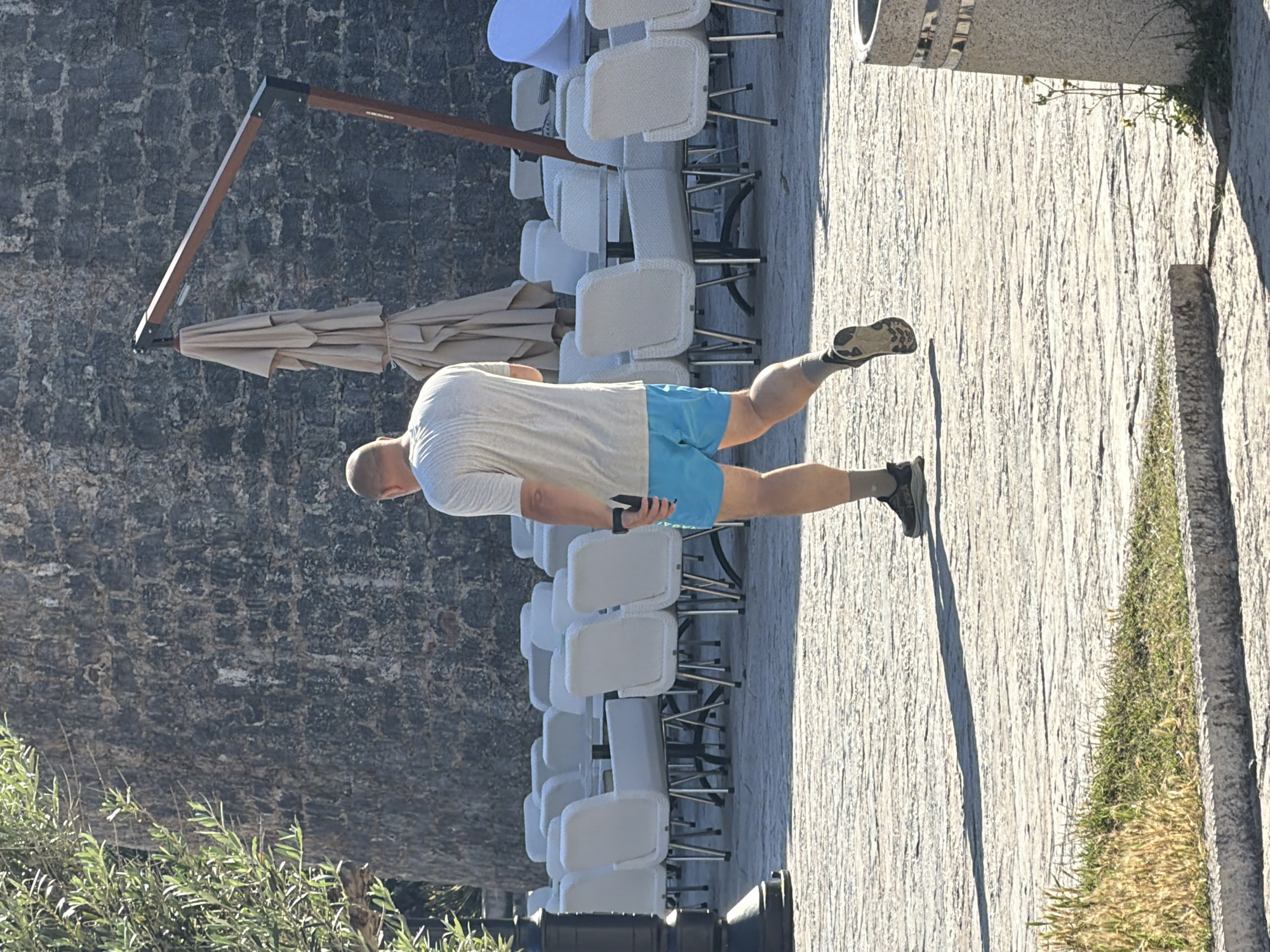 A bald man with a light-colored t-shirt and bright blue shorts walks away from the camera. He is on a stone-paved area that appears to be an outdoor seating area for a cafe or restaurant, with many empty white chairs and tables visible in the background. Behind the seating area is a large, dark stone wall. A large, folded umbrella with a wooden support arm is also visible. To the left of the man and the seating area, there is an olive tree. The scene is lit by sunlight, casting shadows on the paved ground. The location is the outdoor seating area of a cafe in the old town of Budva, Montenegro, near the city walls.