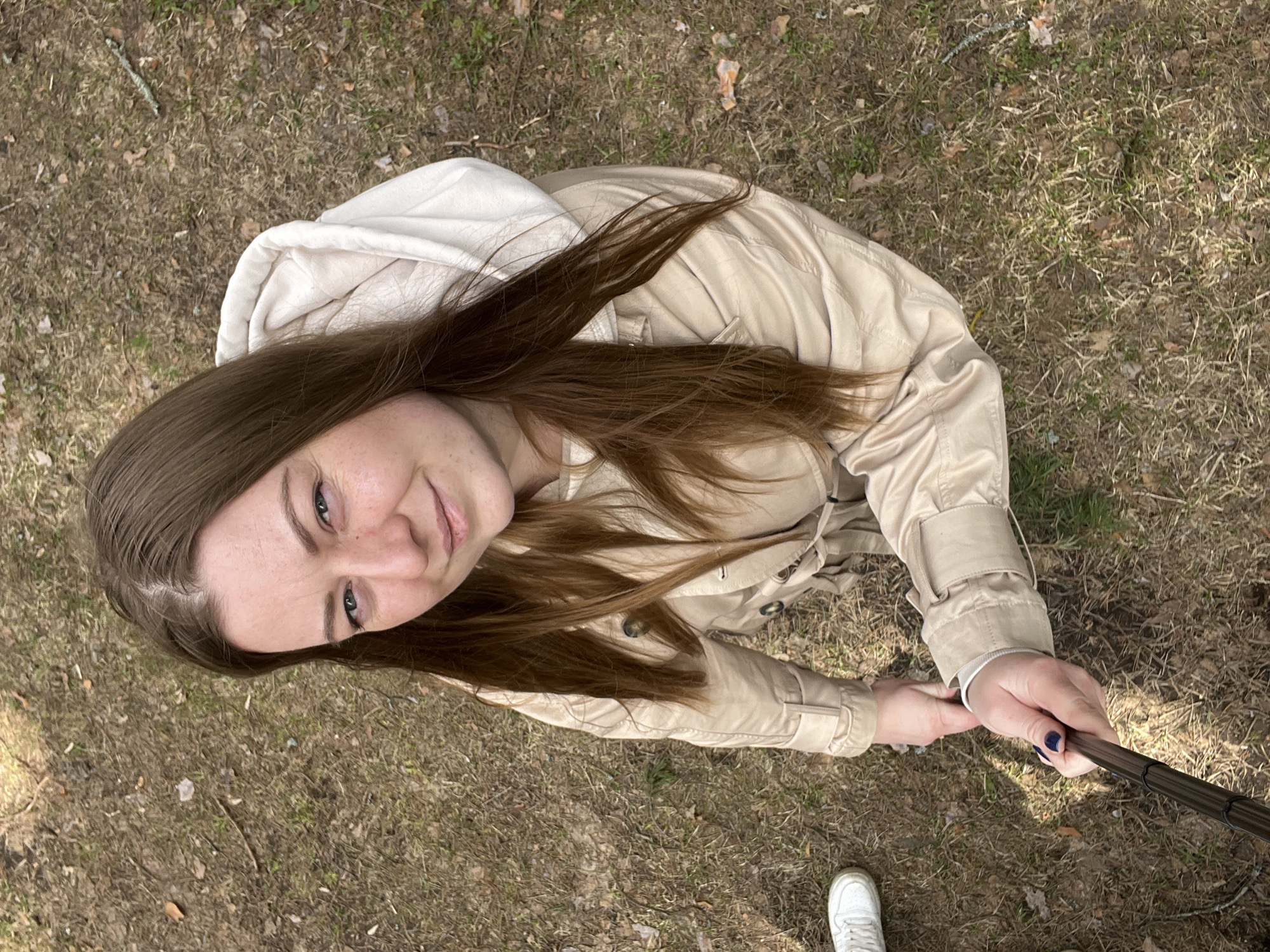 A woman is photographed from above in Razdolye, Russia. She is holding a selfie stick. She is wearing a light beige trench coat over a white hooded sweatshirt. She has long brown hair and is smiling slightly. She is standing on a ground covered in dry grass and leaves. A single white sneaker is visible in the bottom corner of the frame.
