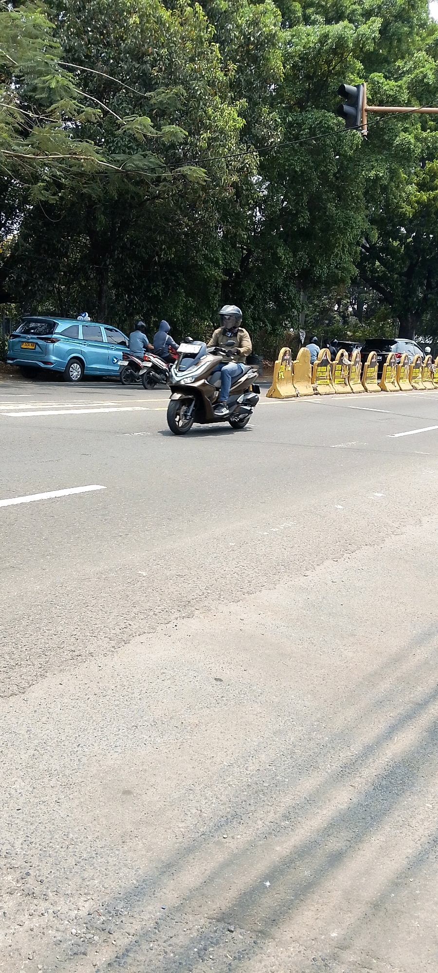 A person is riding a scooter down a street in Depok, Indonesia. The person is wearing a helmet and a light brown jacket. The scooter is light brown. In the background, there is a light blue car, two motorcycles, and several yellow traffic barriers. The trees are green, and the sky is a bright white. The road is paved with asphalt and has white lane markings. A traffic light is visible in the upper right corner of the image.