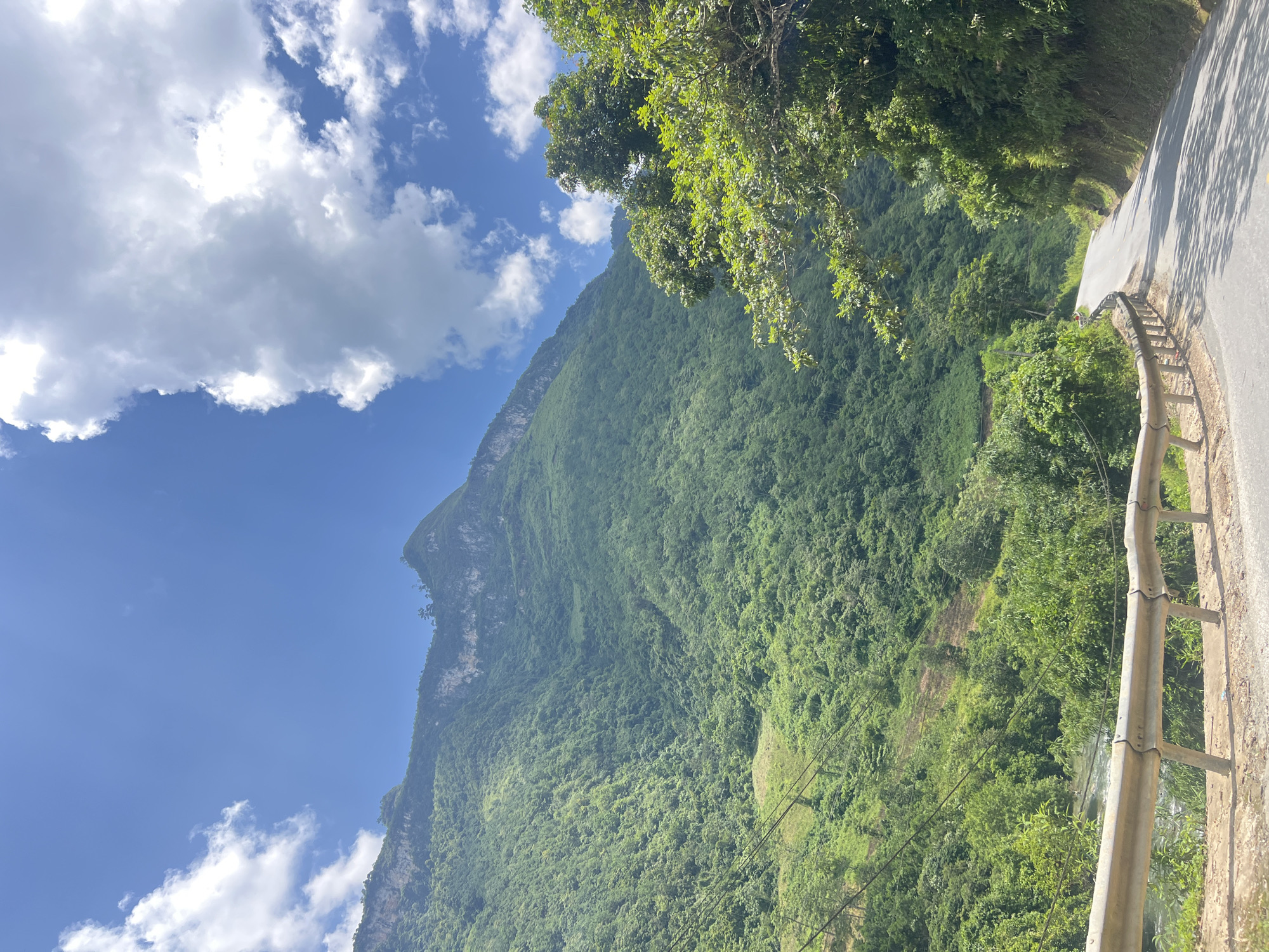 In the city of Minh Tân Commune, Vietnam, a winding asphalt road curves through a lush, mountainous landscape under a bright blue sky dotted with white clouds. The road is bordered on the right by dense, green trees and foliage, casting shadows onto the asphalt. On the left, a metal guardrail separates the road from a steep embankment that drops down to a flowing river. The mountainside is covered in thick, green vegetation, with some areas appearing to be terraced farmland. The scene conveys a sense of natural beauty and a remote, rural setting.