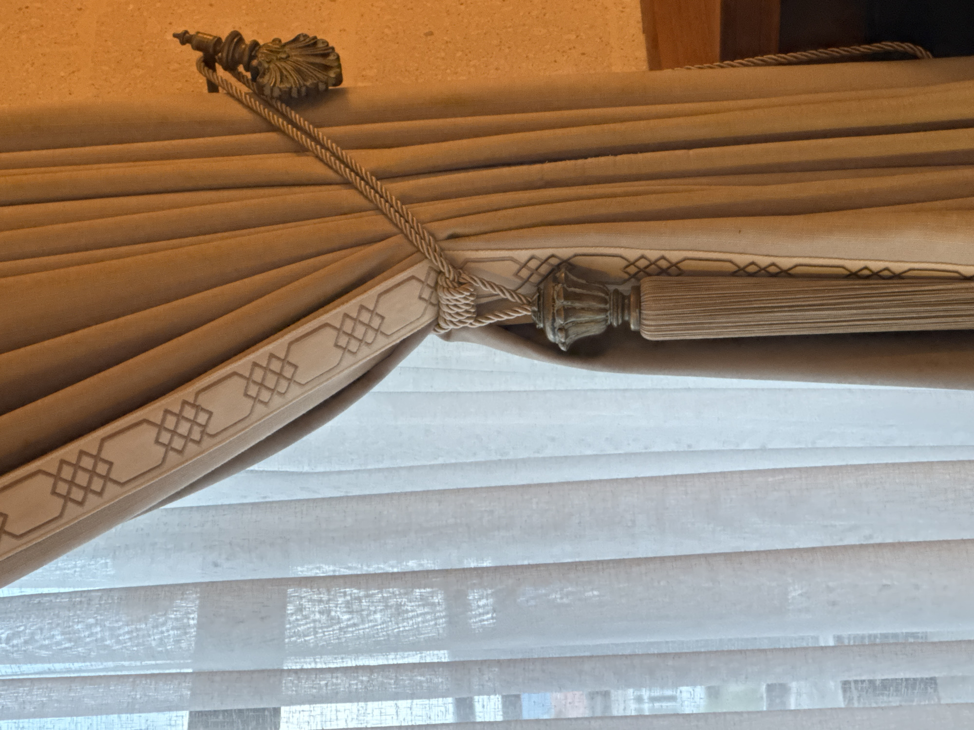 A close-up shot reveals the upper portion of a window treatment, showcasing a beige curtain with a decorative trim featuring a geometric pattern. The curtain is secured by a braided rope that loops around a tassel. Behind the curtain, sheer horizontal blinds filter the light. The location in the city of Ürgüp, Turkey is not discernible from this image.