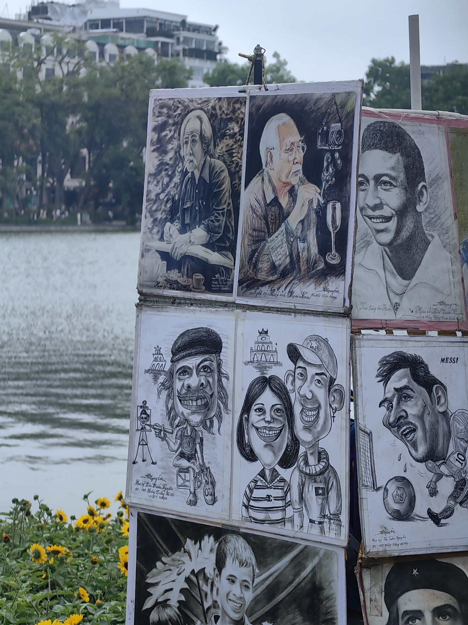 The image shows a display of artwork in Hà Nội, Vietnam, likely near Hoàn Kiếm Lake. The artwork includes portraits and caricatures. Some depict recognizable figures, including footballers Pelé and Lionel Messi. Other caricatures appear to be of tourists. The artwork is signed by various artists, including A. Nguyễn, who provides an address of 128 Hoàng Hoa Thám, Hà Nội, Việt Nam. One piece is signed by "Nghệ sỹ Nhiếp ảnh Moại Nam Hà Nội Việt Nam," indicating the artist is a photographer from Hà Nội. Another artist's signature reads "Họa sỹ Tôn Tuấn Nguyên" with an address of 123 Hàng Trống, Hà Nội. In the background is the lake and buildings in the city. In the foreground, there are blooming yellow flowers.