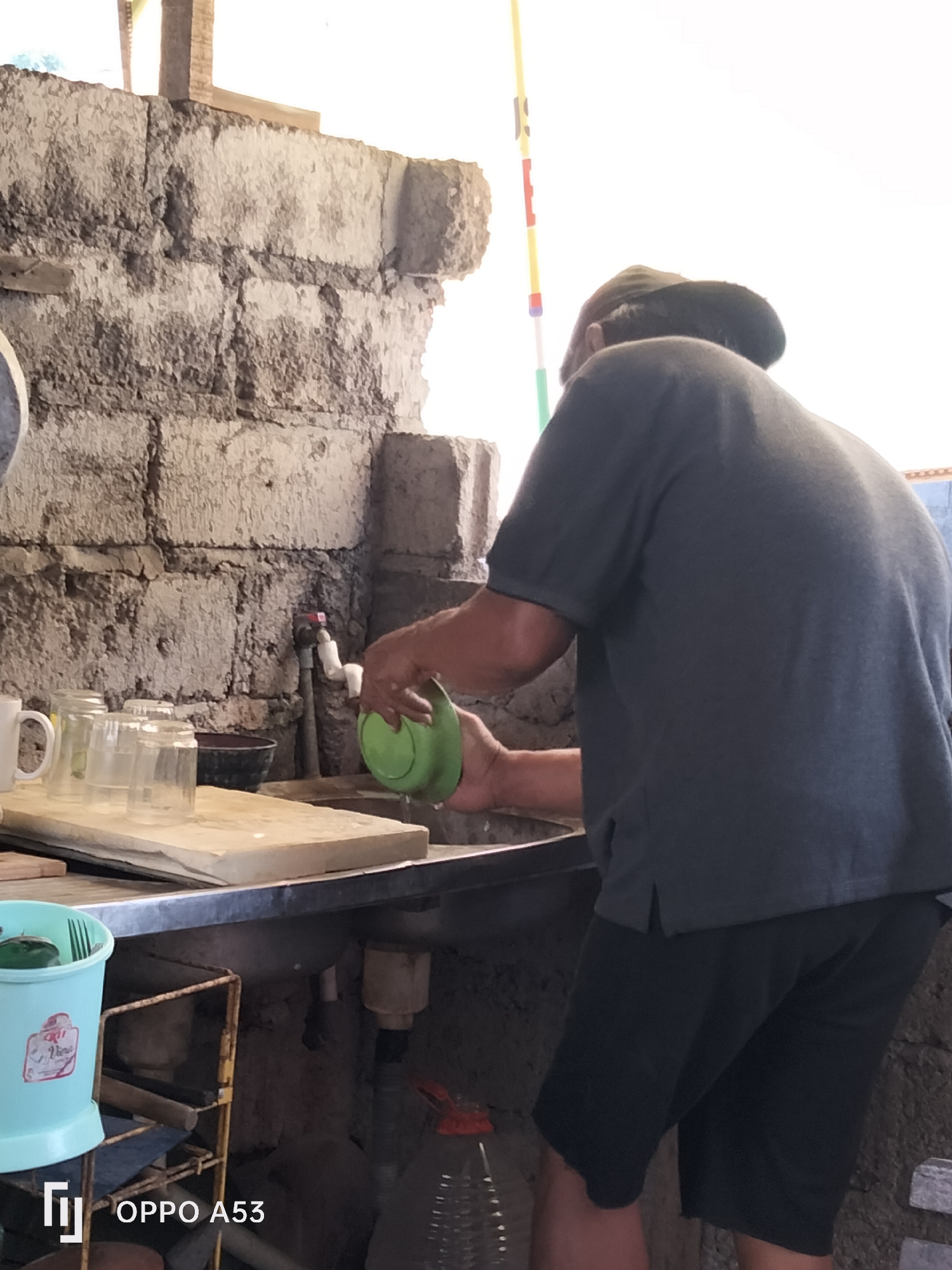 A person wearing a dark gray t-shirt and black shorts washes a green plate in a sink, in an outdoor kitchen with a stone wall. The sink is stainless steel, and a faucet is visible above it. To the left of the sink, there are several clear glasses and a white mug on a wooden cutting board. A light blue plastic container with a handle is on the left side of the image, and a metal shelf with tools is behind it. The background shows a rough stone wall and a bright sky. The location is identified as Denpasar, Indonesia, with an "OPPO A53" watermark at the bottom left of the image.