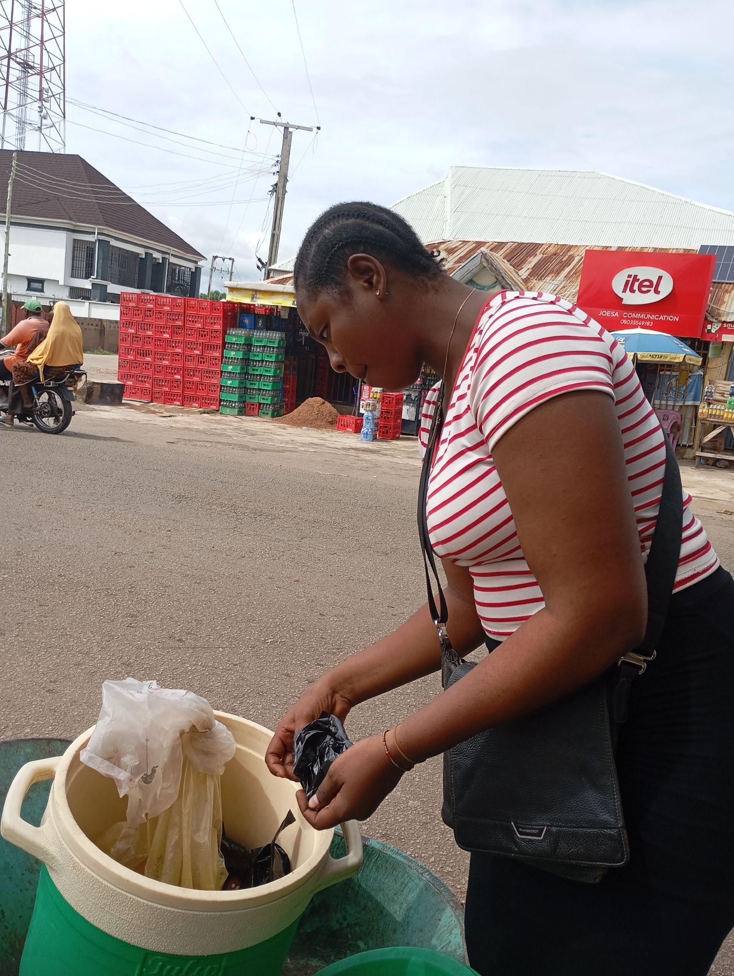 In Shendam, Nigeria, a woman with braided hair, wearing a red and white striped shirt and a black skirt, is seen standing beside a large plastic cooler filled with plastic bags. She is holding a black plastic bag in her right hand, and her left hand is inside the cooler. A black shoulder bag is slung across her body. In the background, there are stacked crates of drinks, a telecommunications tower, a building, and a dirt pile. A motorcycle carrying two passengers, one wearing a yellow headscarf, is visible on the left side of the image. The scene appears to be along a paved road.