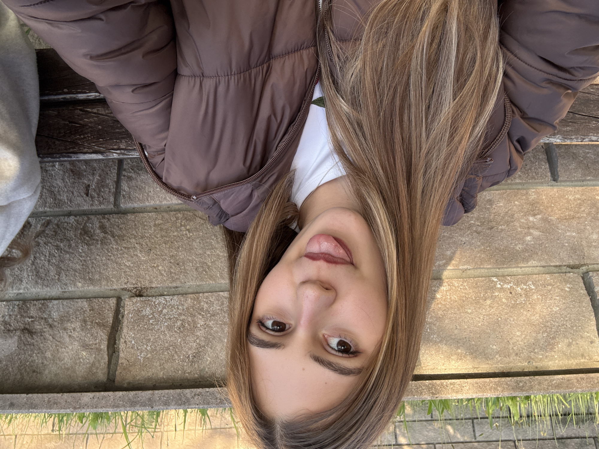 A young woman with long, light brown hair, wearing a brown puffer jacket over a white t-shirt, is sticking her tongue out while looking at the camera. The background appears to be a stone ledge or wall. Grass is visible at the bottom of the frame. The image is oriented at an angle. There is no indication the picture was taken in Panagyurishte, Bulgaria.