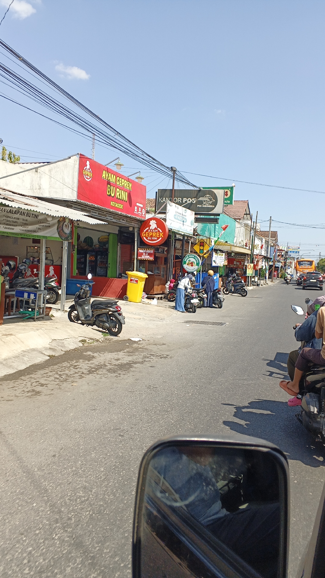 The image captures a street scene in Yogyakarta, Indonesia, likely on a sunny day, with a clear blue sky overhead. The view is taken from a moving vehicle, as evidenced by the reflection in the side mirror and the perspective of the road ahead.
On the left side of the street, there are several small shops and food stalls. One prominent sign reads "AYAM GEPREK BU RINI KOTAGEDE," indicating a fried chicken restaurant. Next to it, another establishment with a sign in Indonesian suggests it sells "lalapan" (vegetables) and other food items. Several motorcycles are parked in front of these shops, and some are on the sidewalk. A bright yellow trash bin is visible.
Further down the street, more businesses line the road. A "KANTOR POS" (Post Office) sign is clearly visible, alongside other commercial establishments. People are seen interacting with motorcycles and walking along the sidewalk. A traffic light is present at an intersection further down the road.
On the right side of the street, the view shows more vehicles, including motorcycles with riders, and cars. A bus can be seen in the distance. Shadows of people and objects are cast on the road, indicating the direction of the sun. The road surface is asphalt, and there are some painted markings. Electrical wires are strung across the top of the scene. The overall impression is of a busy, everyday street in an Indonesian city.