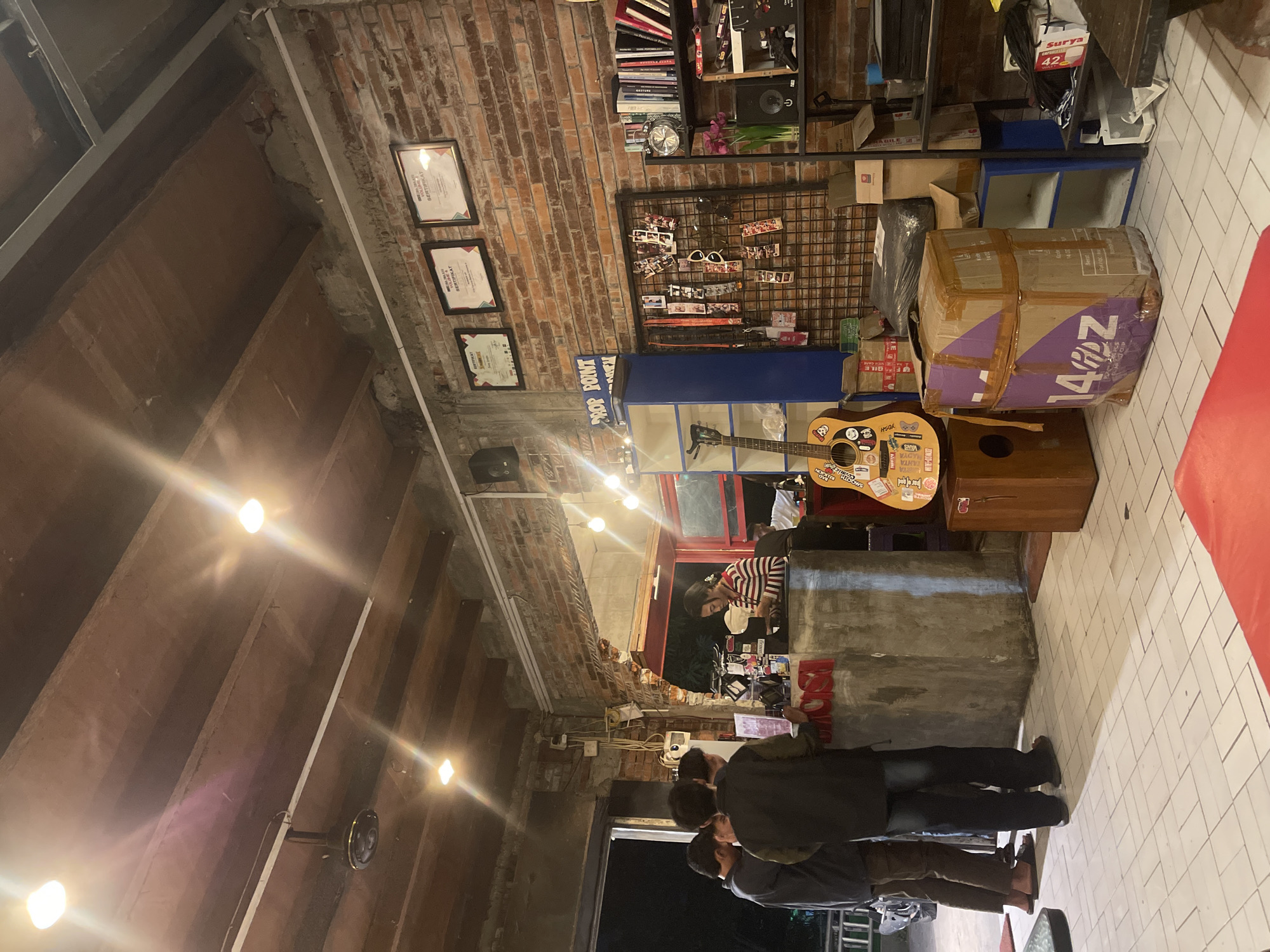 This image depicts a dimly lit indoor scene, likely a shop or cafe, with exposed brick walls and wooden ceilings. In the foreground, a group of four individuals, all appearing to be adult males dressed in casual clothing, are gathered around a concrete counter. Three of them are facing the counter, engaged in an interaction with a person behind it, who appears to be a young woman. She is wearing a striped long-sleeved shirt and is attending to something on the counter. The counter itself has red lettering that is partially obscured, but appears to spell out "MOSI." To the left of the counter, an open doorway leads to an outdoor area with a railing.
To the right of the counter, there is a blue shelving unit with a guitar leaning against it. A wooden box or stool is placed next to the guitar. Further to the right, a black wire rack displays various lanyards and small items. Behind this rack is a bookshelf filled with books, a clock, and a vase with pink flowers. Various boxes, including some taped with purple and brown tape, are stacked on the floor and on shelves throughout the scene. The floor is tiled with white squares. The overall ambiance suggests a casual, possibly bohemian, commercial establishment. The image is identified as being located in the city of Pagerwojo, Indonesia.