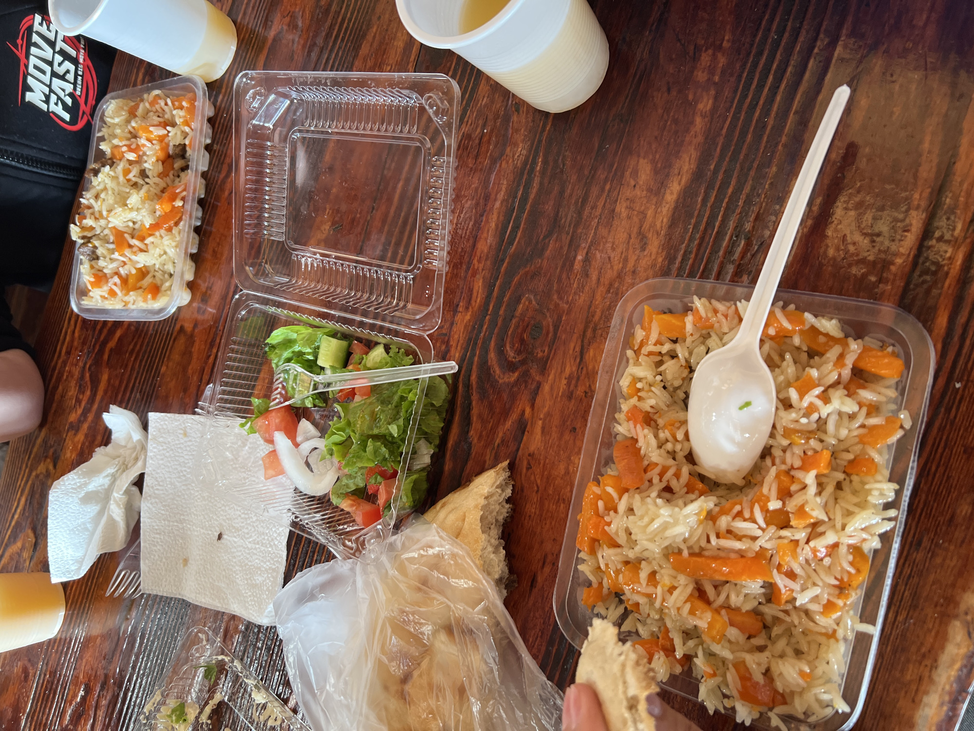 The image depicts a wooden table at a restaurant in Listvyanka, Russia, with several containers of food and drinks. A close-up shot reveals two plastic containers of food, one containing rice with carrots and meat, and another with a salad of lettuce, tomatoes, cucumbers, and onions. There is also a plastic cup with a beverage, and a piece of bread in a plastic bag. A person's hand is visible in the lower left corner, holding a piece of the bread. The overall scene suggests a casual meal setting.