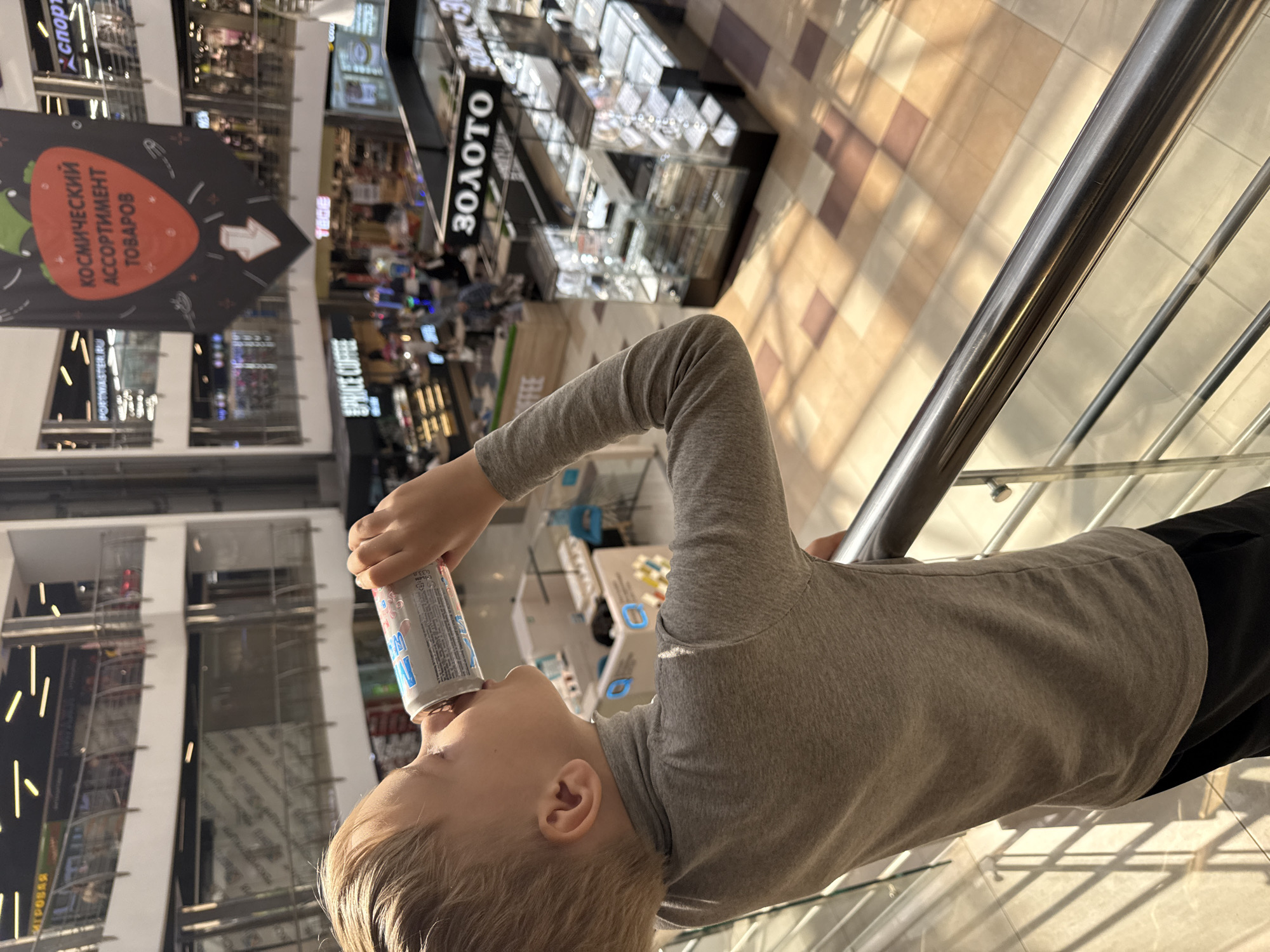 A child is drinking from a can in a mall. The mall appears to be located in Dmitrov, Russia, based on the Russian text visible on a nearby sign. The child is wearing a gray long-sleeved shirt and black pants. The child is standing near a railing, and the can is elevated toward the child's mouth. The mall interior is multi-level, with various retail shops and kiosks visible in the background. One visible sign reads "Золото" (Gold) and another says "Космический Ассортимент Товаров" (Cosmic Assortment of Goods). The floor is tiled in a checkered pattern of light and dark colors.