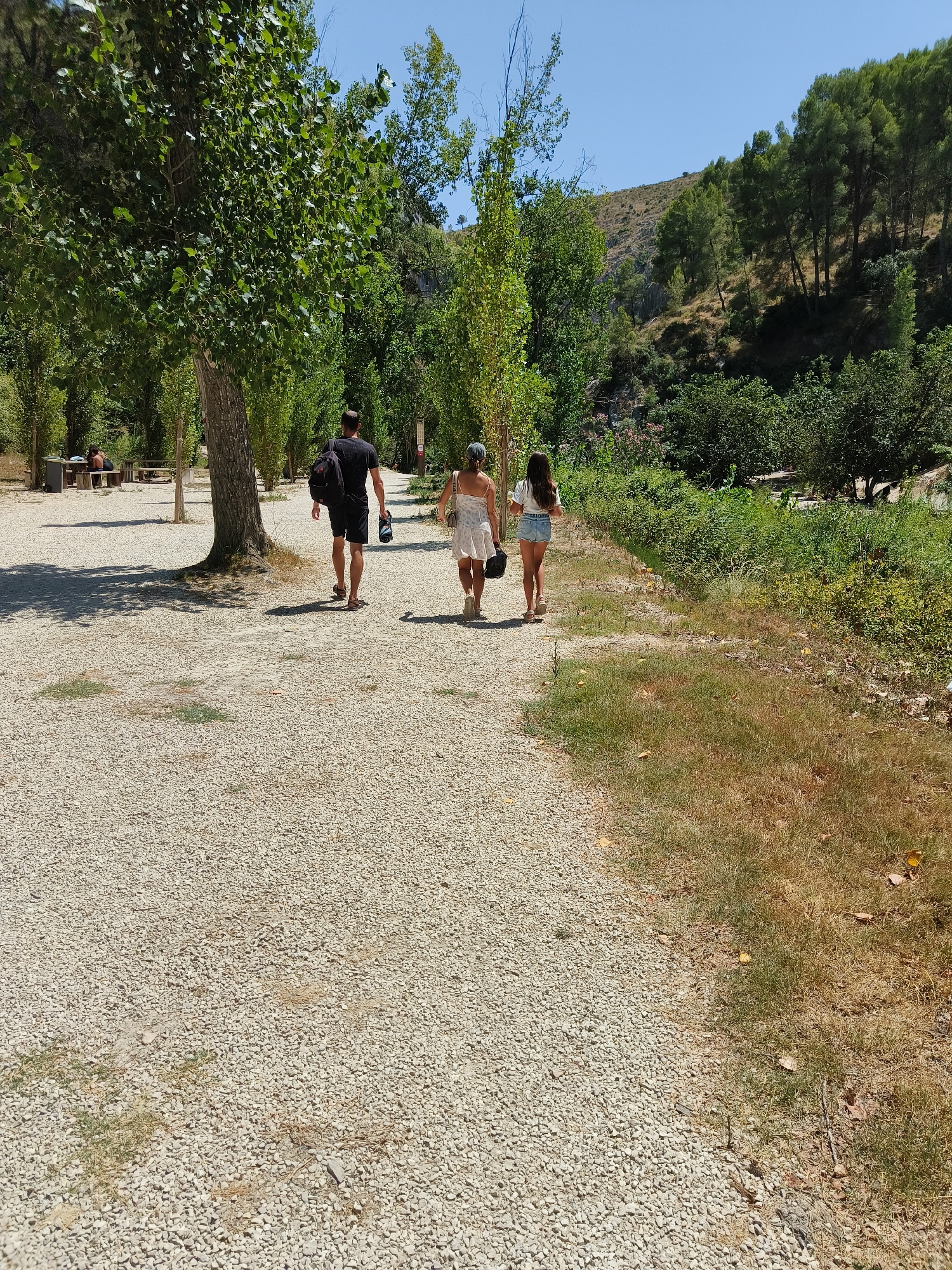 The image shows a sun-drenched scene with a gravel path winding through a park-like area in Ontinyent, Spain. Three individuals are walking away from the camera on the path. The person in front is a woman wearing a white dress and carrying a dark bag. Beside her is a younger woman in denim shorts and a white t-shirt, also holding a dark bag. Following behind them is a man in a black t-shirt and shorts, carrying a black backpack.
The path is lined with trees, some of which cast dappled shadows on the ground. To the right of the path, dry grass and vegetation are visible, suggesting a warm, dry climate. In the background, a lush green hillside with scattered trees rises under a clear blue sky. Further back, there is a darker area of dense trees and foliage, possibly indicating a more natural, less manicured area. To the left, in the distance, some picnic tables and benches can be seen under trees, with two people seated. The overall impression is of a pleasant, outdoor recreational space.