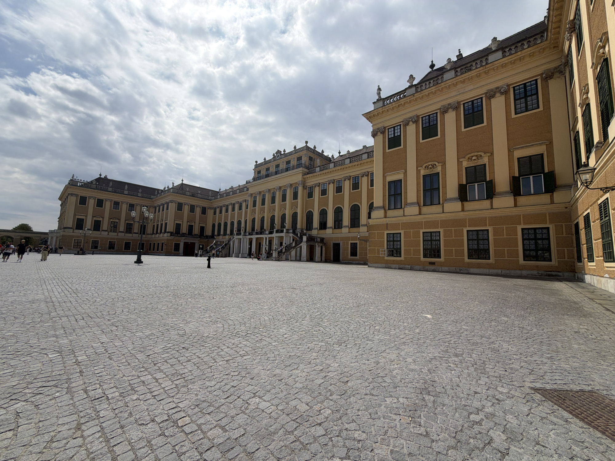 The image depicts a wide, paved courtyard in front of a grand yellow palace, likely Schönbrunn Palace in Vienna, Austria. The palace facade, characterized by its classical architecture, features numerous arched windows with dark shutters and ornate decorative elements. Several individuals are present in the courtyard, some walking and others standing. The ground is paved with cobblestones arranged in a radiating pattern. The sky is partially clouded with a mix of light and dark clouds. The scene conveys a sense of historical grandeur and public space.