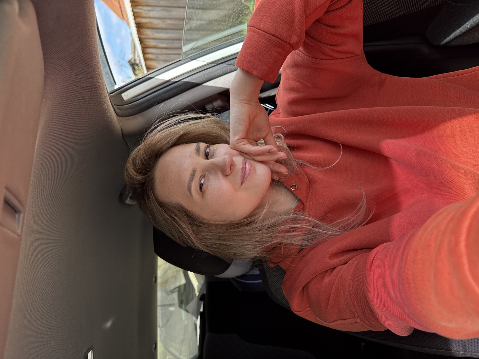 A young woman is sitting in the driver's seat of a car, turned to face the camera for a selfie. She is wearing an orange hoodie and has light blonde hair. Her right hand is lightly touching her face. The car window is partially open, revealing the exterior, which includes a wooden structure possibly a house or shed, and a glimpse of a blue sky. The interior of the car is visible, showing elements like the ceiling, the car seat, and parts of the dashboard. The location is Chaikovsky, Russia.