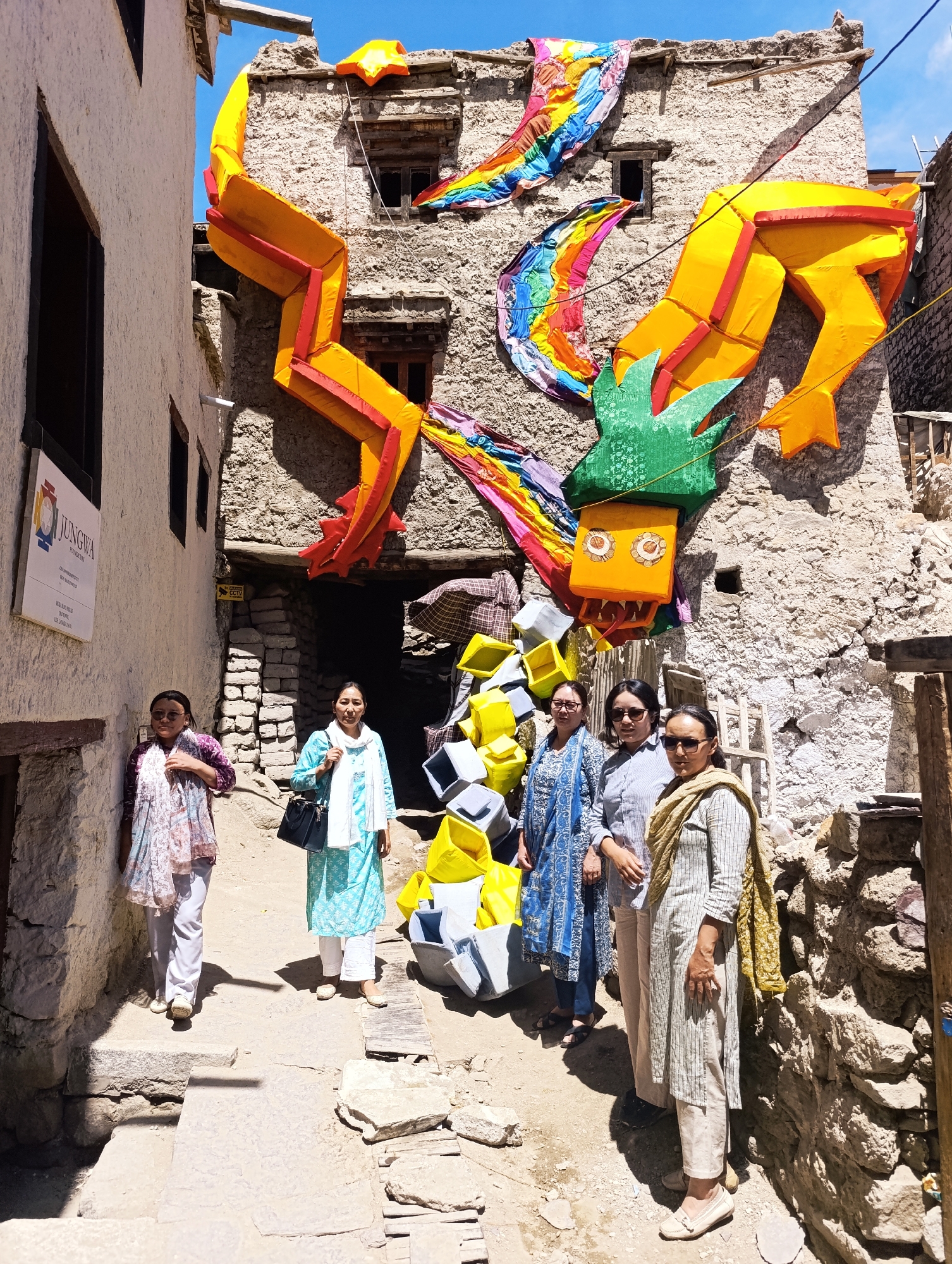 In the city of Unknown, Unknown, a bright yellow dragon decoration adorns the exterior of a stone building. The dragon, with red accents and green spikes, appears to be made of fabric and is draped across the facade. Below the dragon, a group of five women stand in a narrow, dusty alleyway. The women are dressed in traditional clothing, with various colors and patterns. Some wear scarves and head coverings. In front of them, a pile of yellow and grey rectangular objects are stacked, possibly for an event or display. The building behind the women is made of rough stone, with wooden beams and small windows. The ground is uneven and covered with dirt and stones. A white sign with blue and red text is visible on the left side of the alley. The scene is illuminated by natural daylight, casting shadows on the ground.