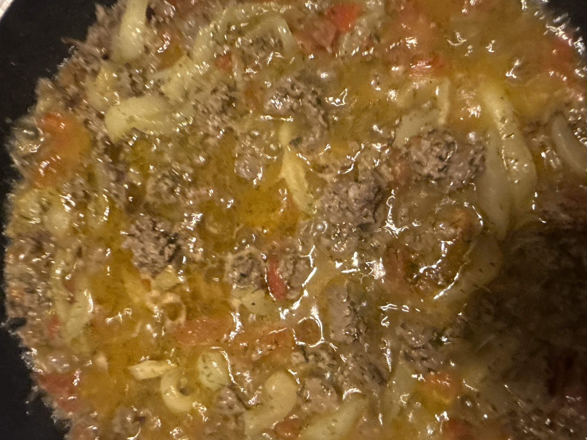 This image shows a close-up, overhead view of a meal cooking in a dark pan. The dish appears to be a pasta or rice-based casserole with ground meat, chunks of tomato, and possibly onions or other vegetables in a rich, golden sauce. The surface of the liquid is shimmering and reflects light, indicating it is hot and possibly simmering or boiling. The textures are varied, with the coarse grain of the meat contrasting with the smoother sauce and softer vegetables. The overall impression is one of a hearty, home-cooked meal in progress. The location of this scene is Unknown, Unknown.